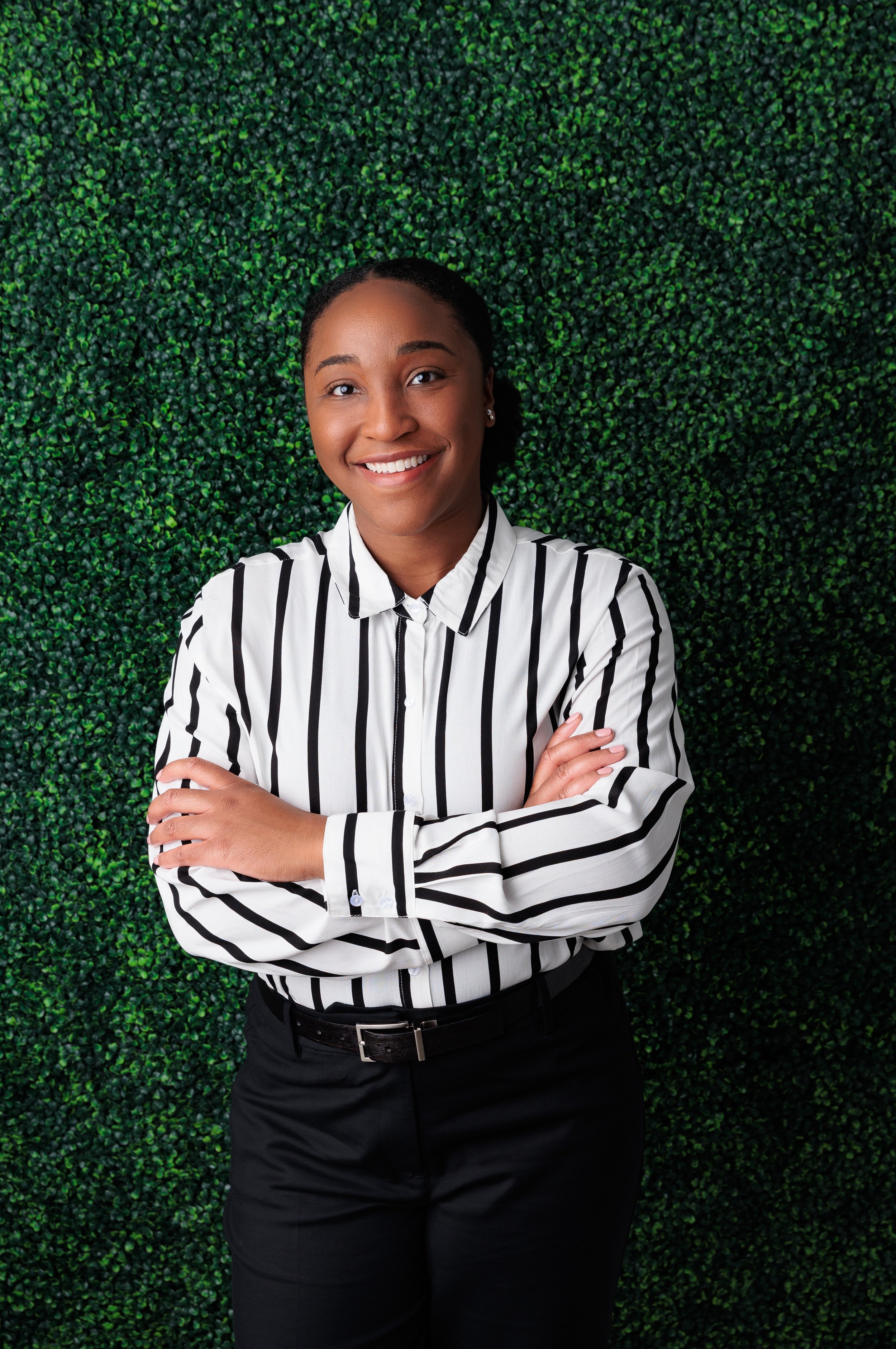 Kelsey who is a black female smiles at the camera with her hair pulled back, swearing a black and white striped blouse and standing in front of a grass wall
