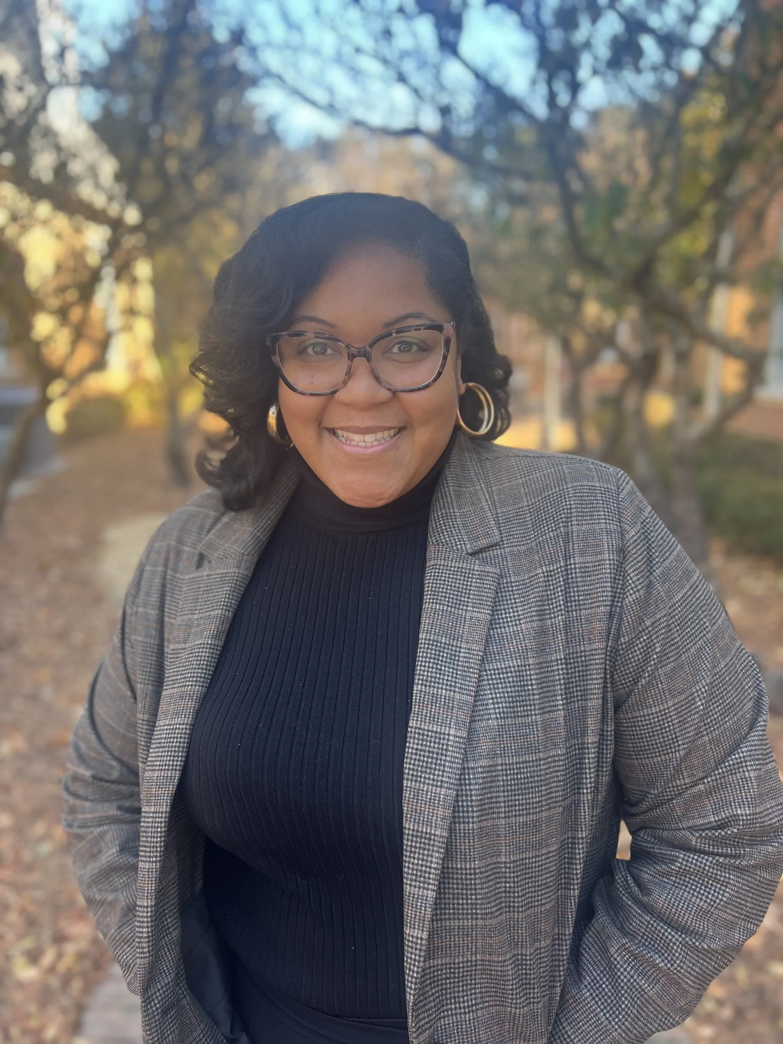 An image of Bianca Mason who is a black female, she wears glasses, a black shirt and pinstripe blazer and stands outside.