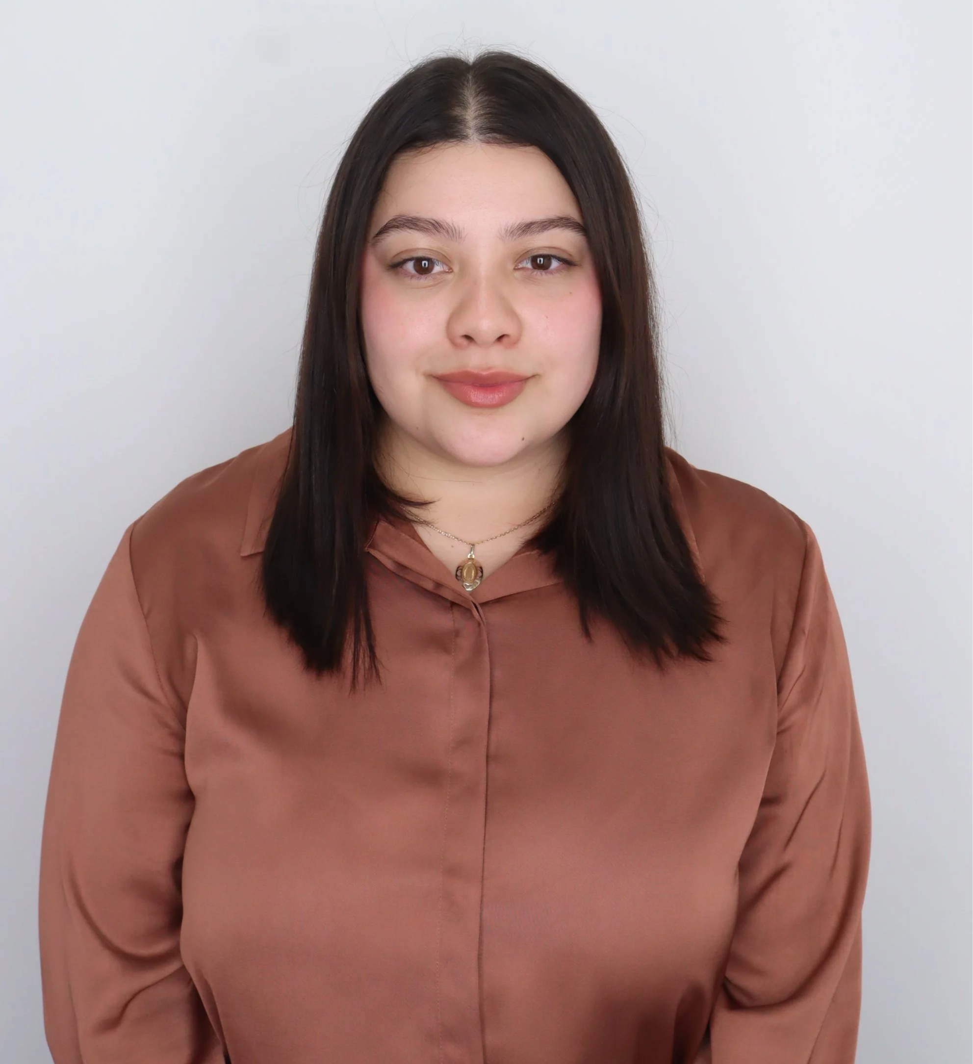 Jennifer, a hispanic female with dark hair past her shoulders and a brown blouse stands in front of a blank wall and smiles softly at the camera