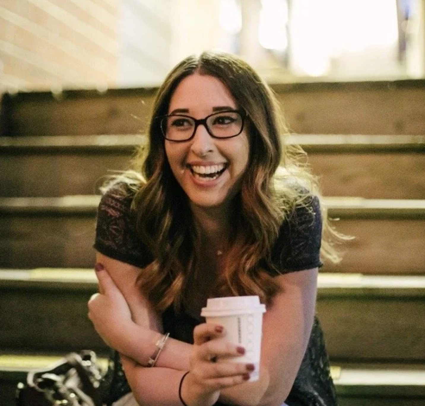 an image of Eden who is white, brunette hair, smiling with her hair down, siting on a staircase and holding a coffee cup. She wears glasses and a black shortsleeve shirt
