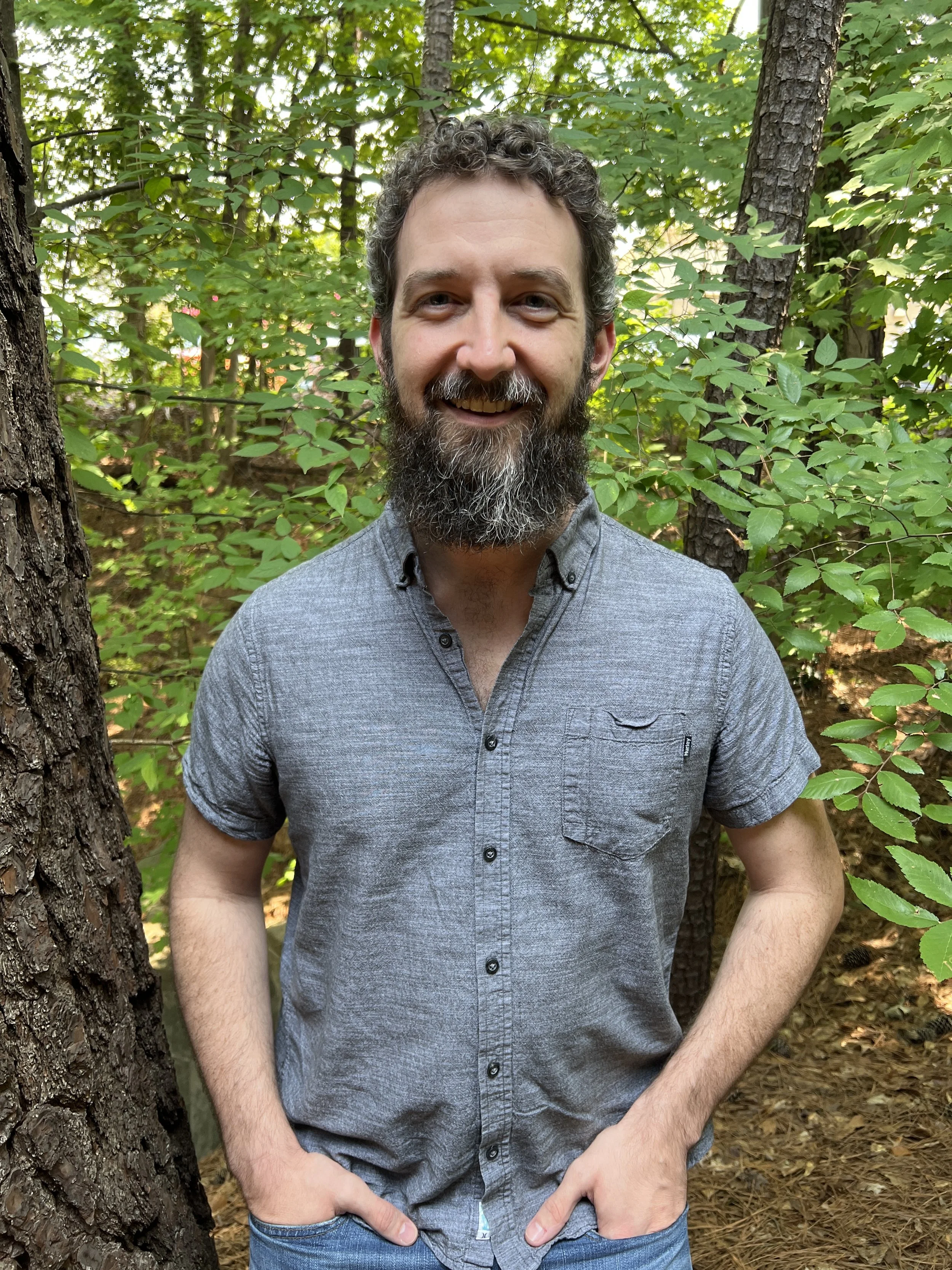 An image of Zack who stands outside, smiling at the camera. He wears a shortsleeve gray button down, is a white male with a salt & pepper beard.