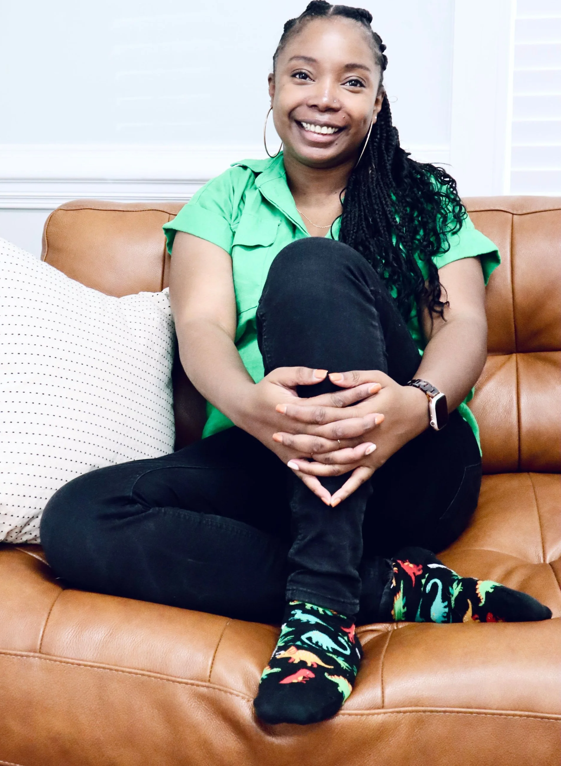 An image of Erica who is a black female, she has braids is wearing a green blouse, black jeans, and dinosaur socks. She sits on a brown leather couch.