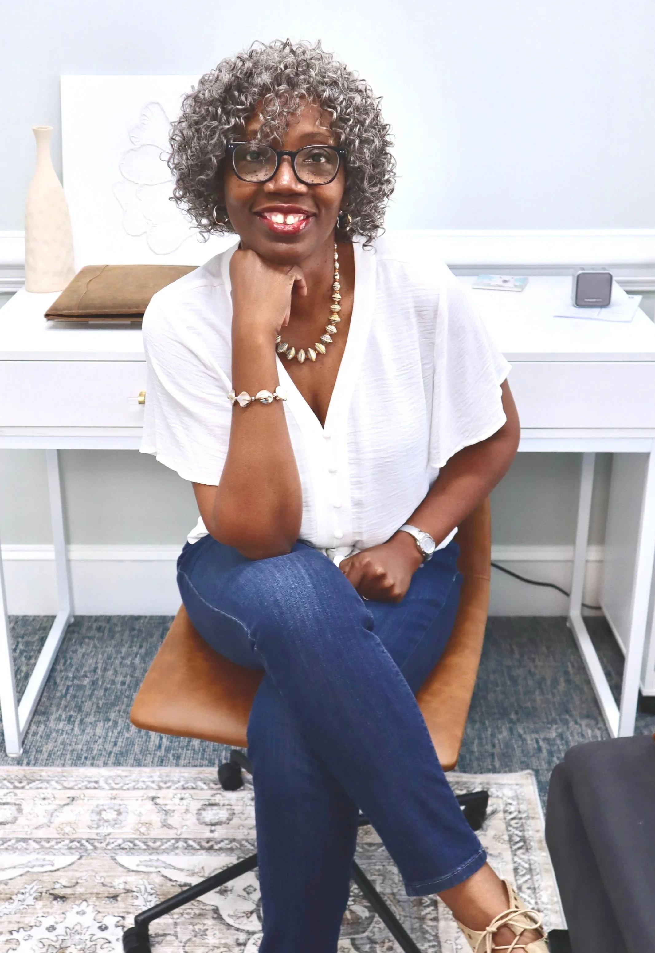 An image of Cassandra who is a black female wearing a white blouse, blue jeans, and sitting in a brown chair. She smiles at the camera and wears glasses.