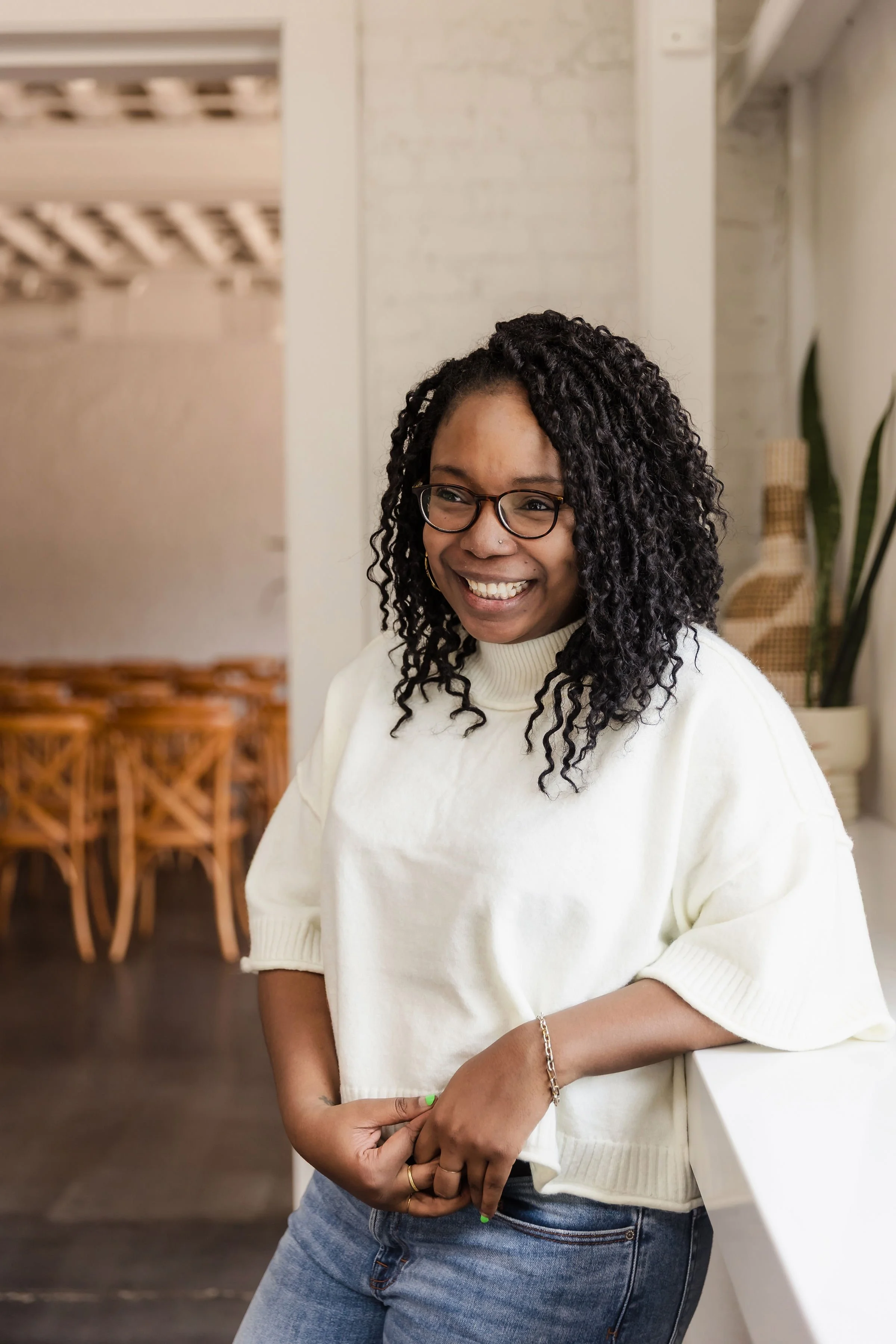 An image of Erica who is black with curly hair and glasses. Smiling while wearing a cream blouse, blue jeans, standing and leaning against a counter