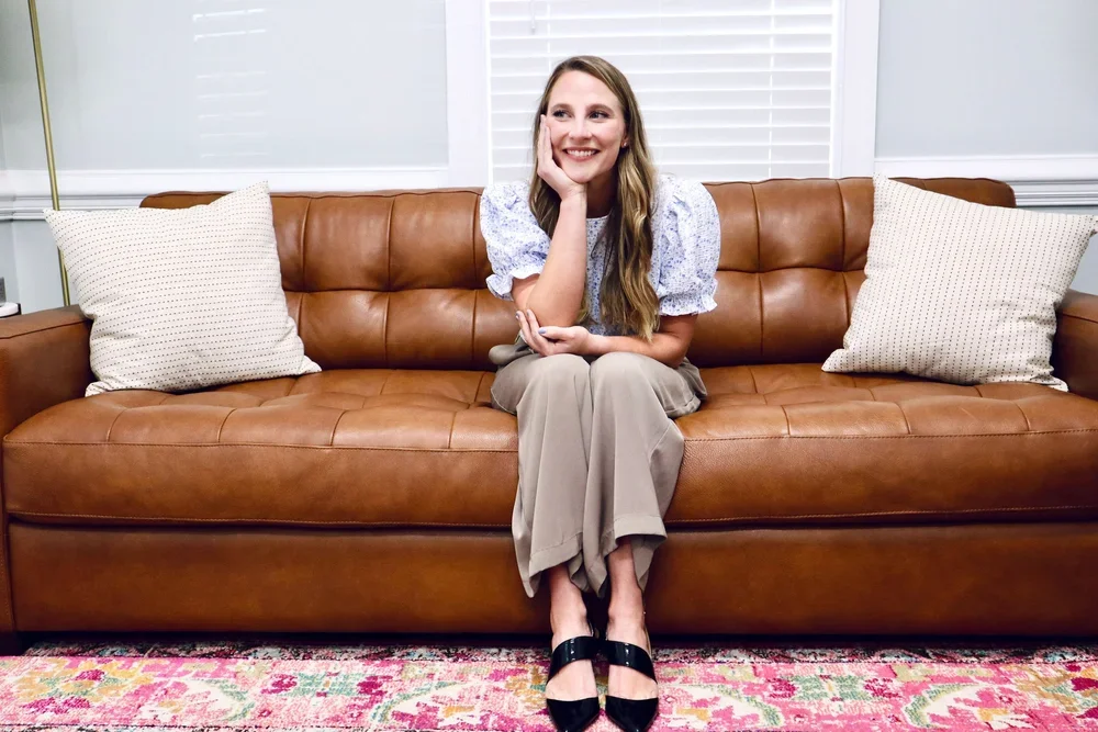 Emily who is a white female is wearing a blouse, khaki pants and smiles at the camera while sitting on a brown leather couch