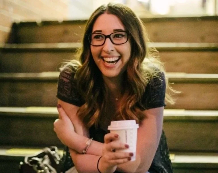 Eden, a white female sits on a staircase holding a coffee cup and smiles at the camera. She wears glasses, and has dark brown hair.