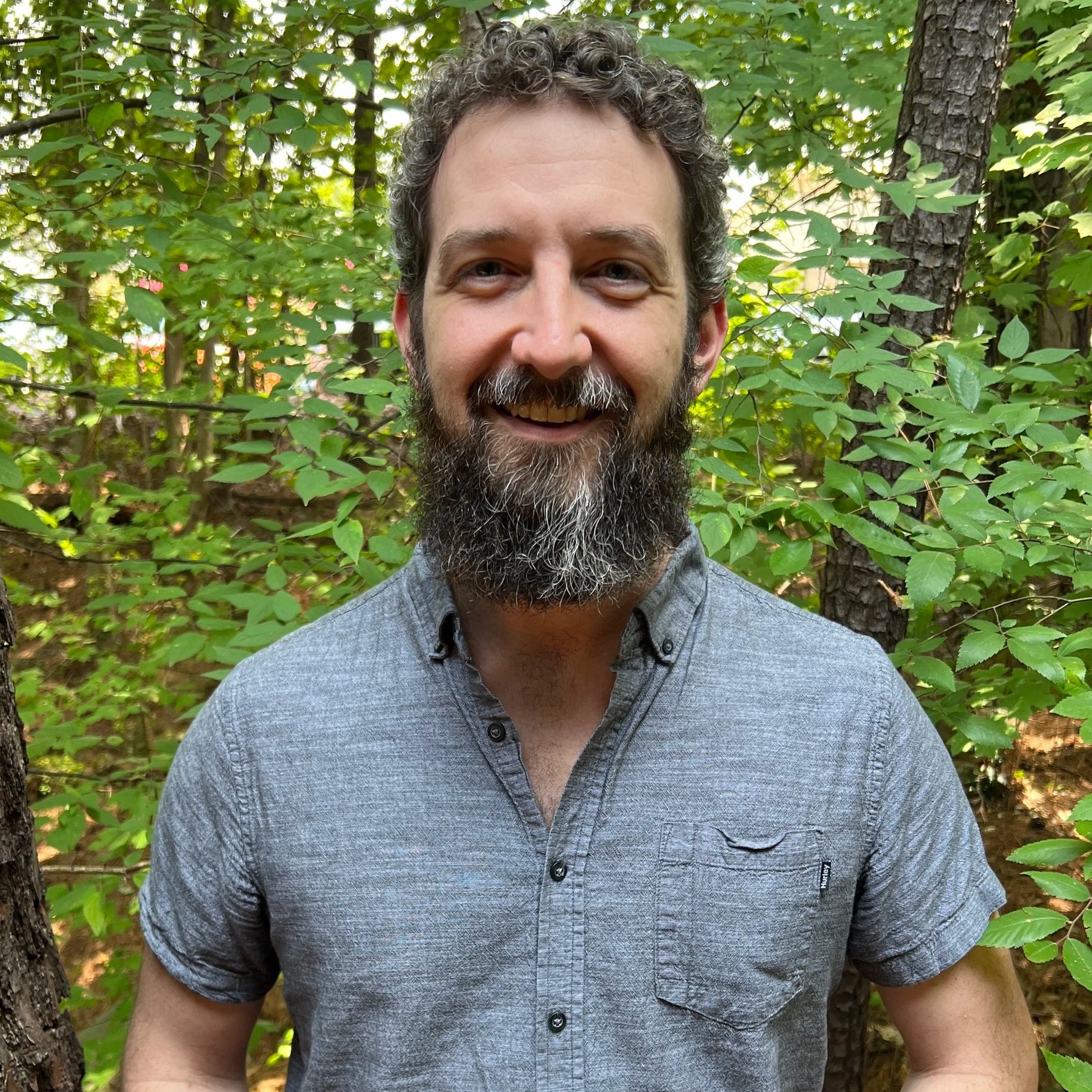 An image of Zack who is a white male, with a salt and pepper beard and he is smiling at the camera. Zack stands outside wearing a grey button down
