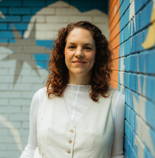 Brianne, a Hispanic & white therapist leans against a colorful wall and smiles at the camera. Her curly hair is down and she is wearing a white and cream blouse/vest