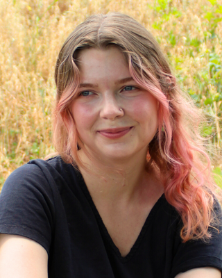 An image of Devin, a white female who sits outside in a black v-neck. She has blonde and pink hair and smiles as she looks off camera