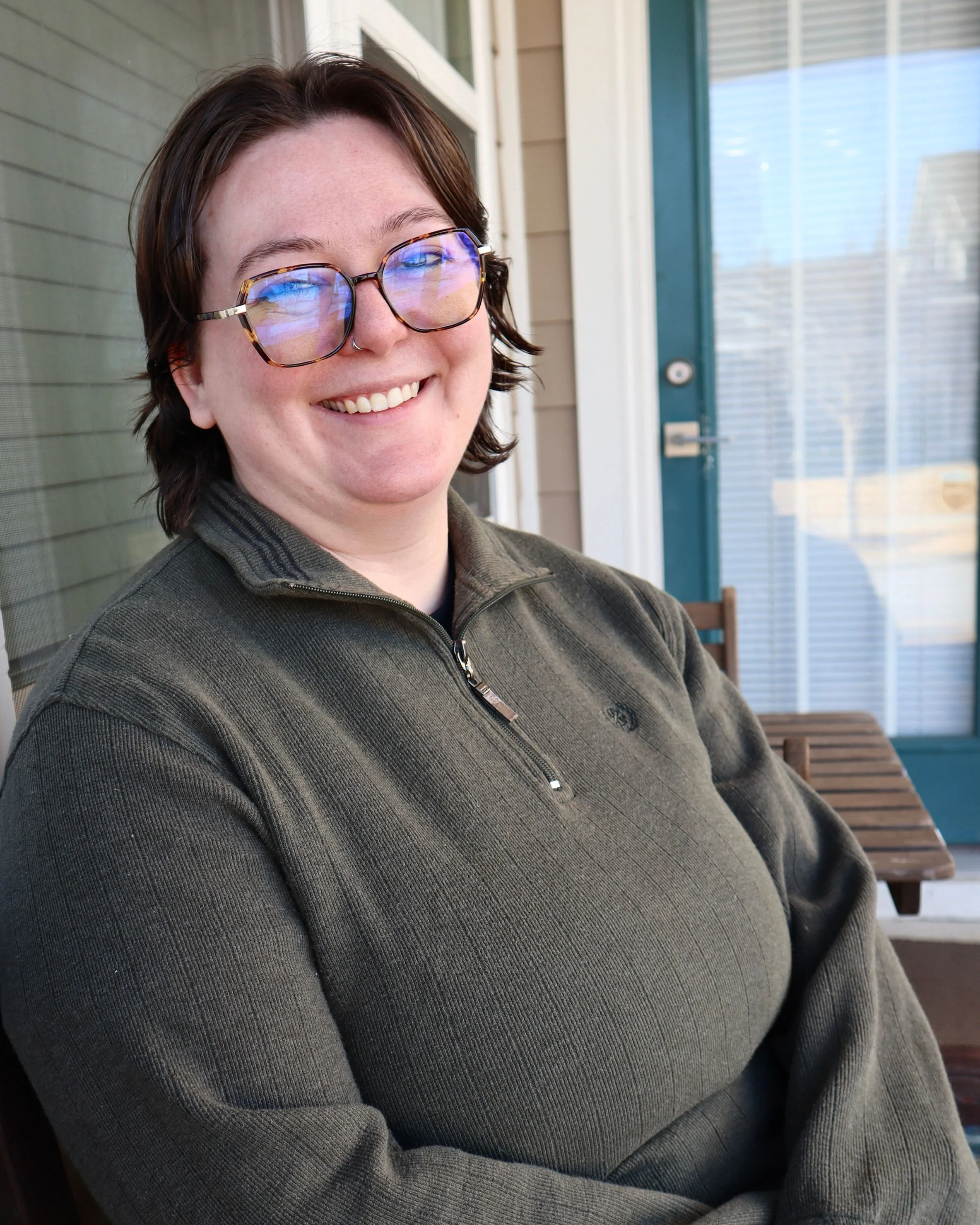 Ria is a white human with brunette hair, wearing a dark green sweater. Ria is wearing glasses, and smiling at the camera.