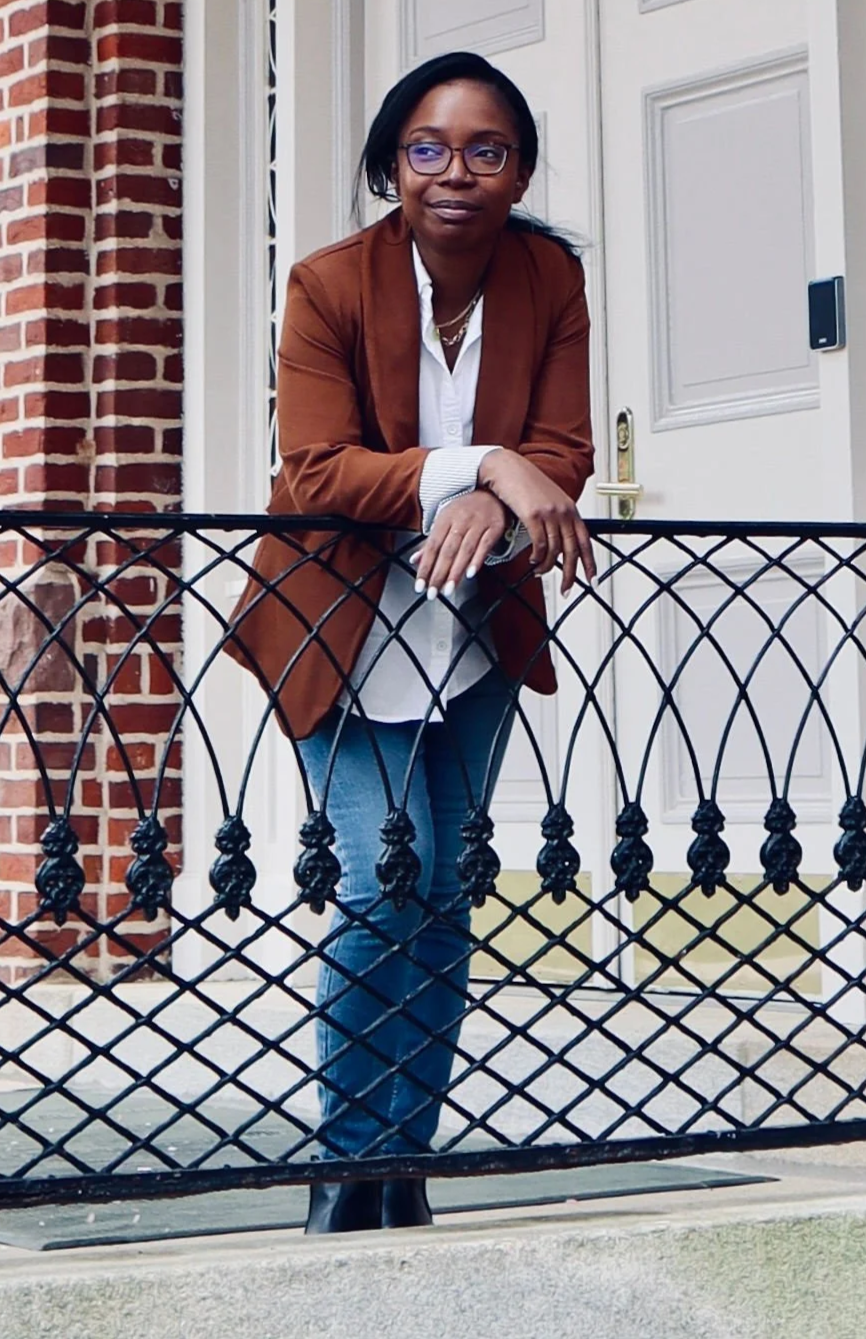 Erica is a black female standing leaning over a balcony wearing jeans a white shirt and brown blazer