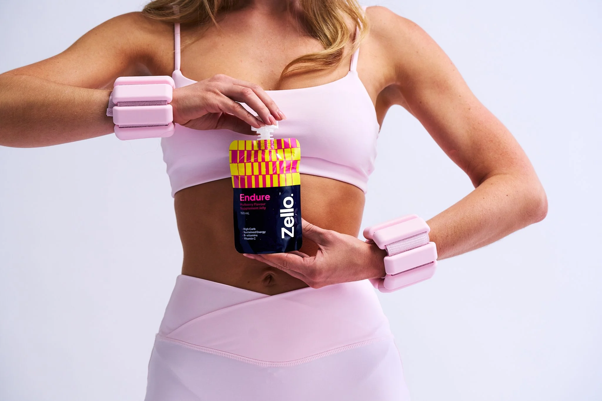 A woman in a pink sports bra and leggings holding a Zelle Endure supplement packet, with pink wellness wristbands on her arms, against a plain background.