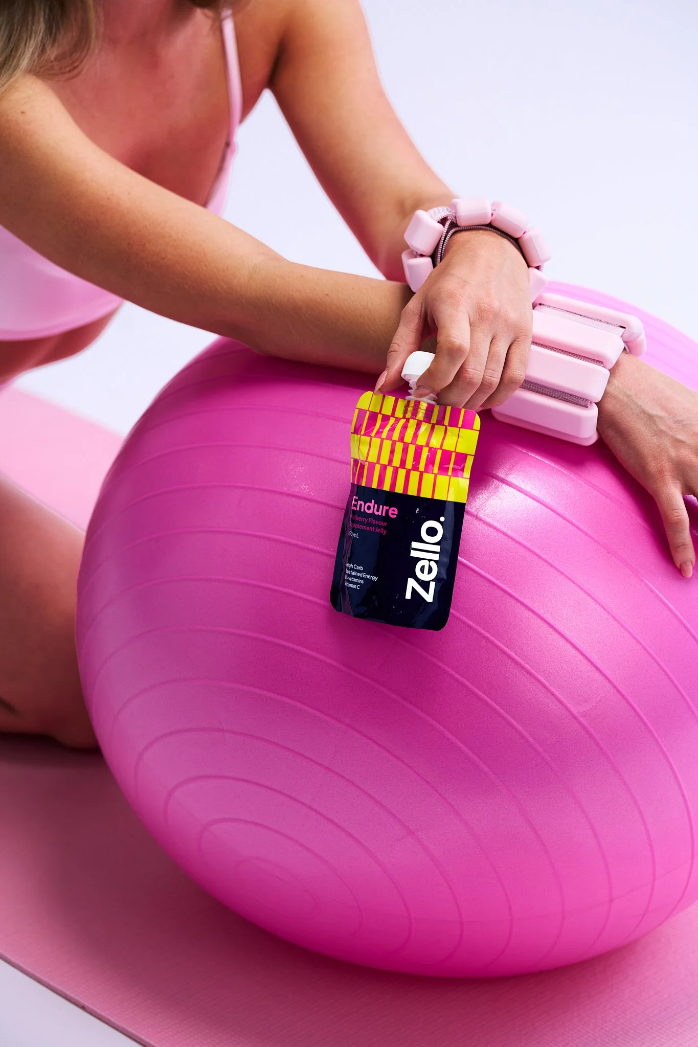 Close-up of a woman wearing a pink tank top and pink leggings, sitting on a pink yoga mat with a pink exercise ball. She is holding a package of pink and purple gummy vitamins.
