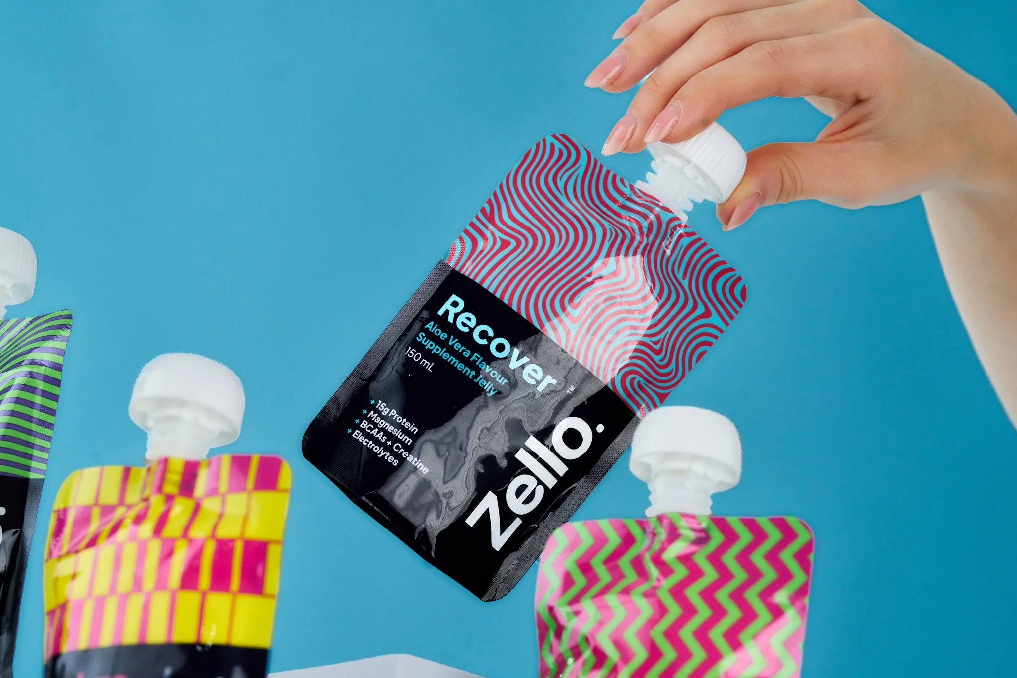 A person is squeezing toothpaste onto a tube of ZeloReviver Aloe Vera flavored jelly supplement amidst colorful, patterned boxes and some white pills on a bright blue background.