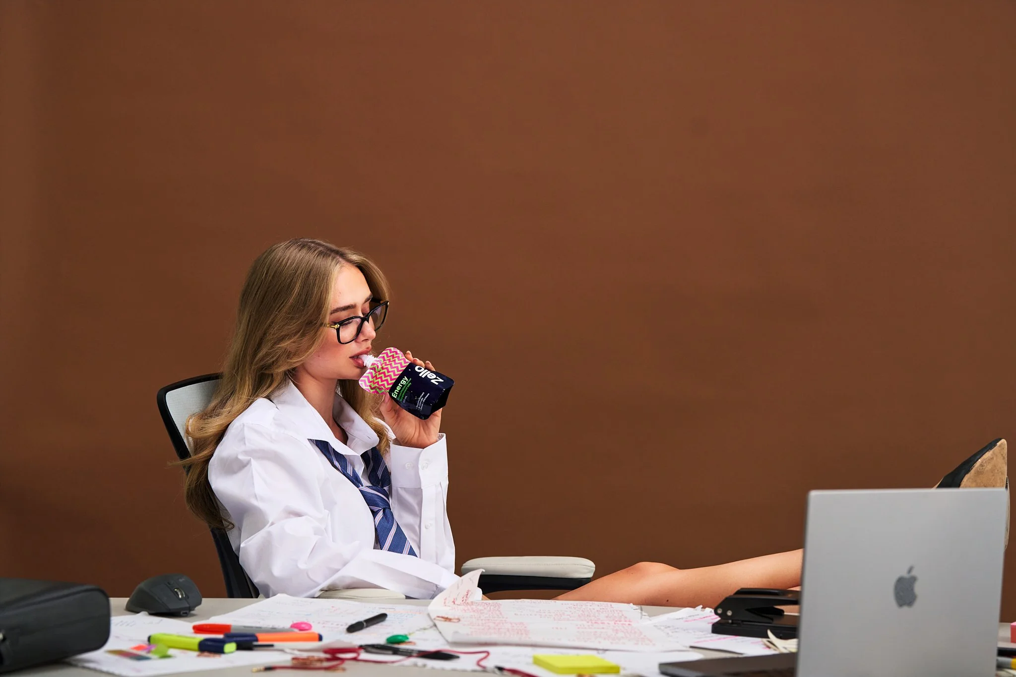 A young woman with long blonde hair, glasses, and a school uniform, sitting at a cluttered desk with papers, pens, and colorful markers, drinking from a small carton of energy drink in an office setting with brown walls.