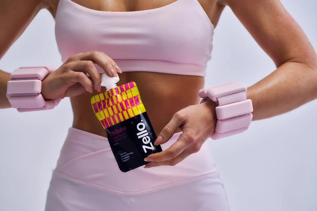 A woman wearing pink workout accessories pours energy gel from a pink and black packet labeled "Endure" into her hand during exercise.