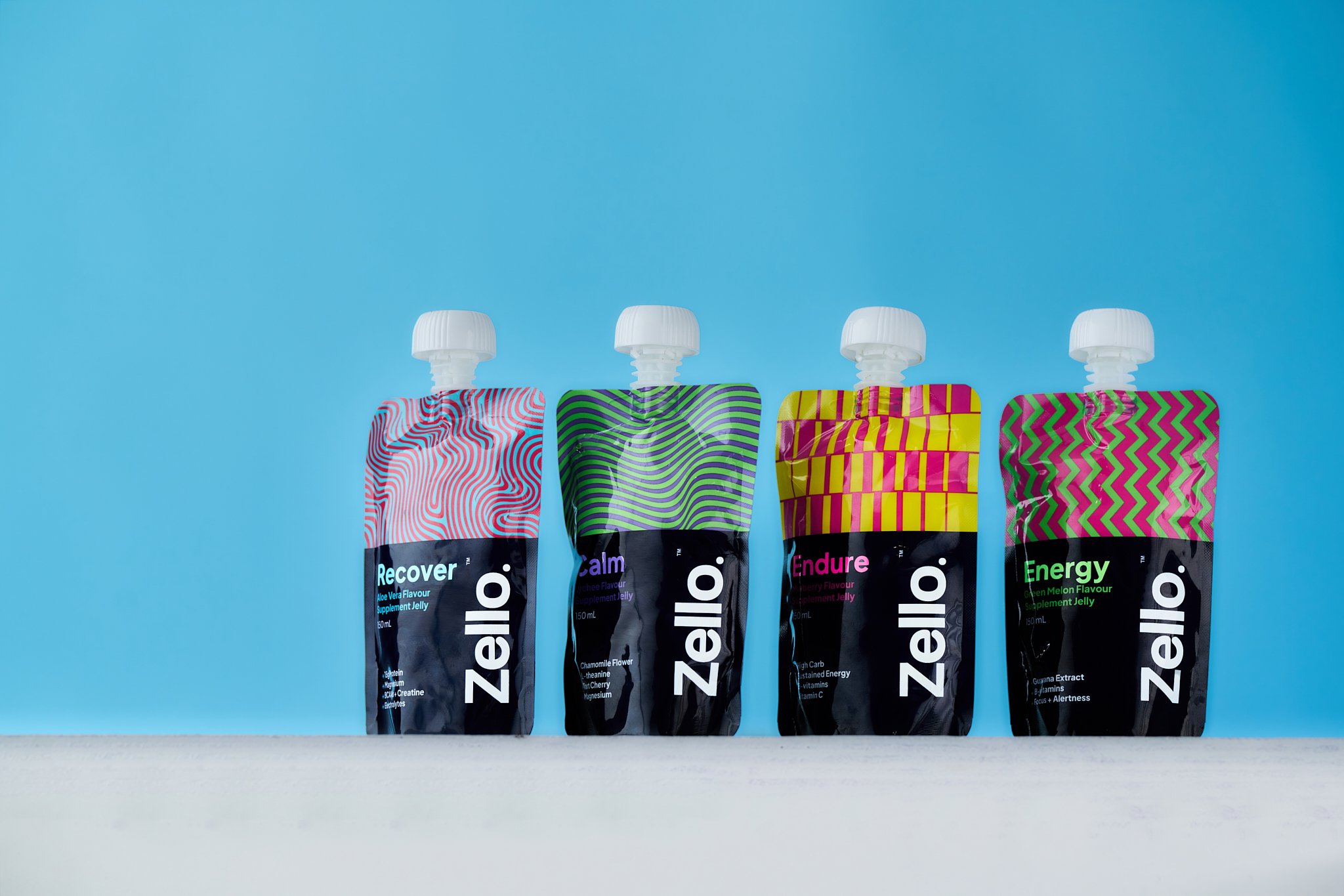 Four colorful gel supplement pouches with twist-off caps lined up on a surface against a blue background.
