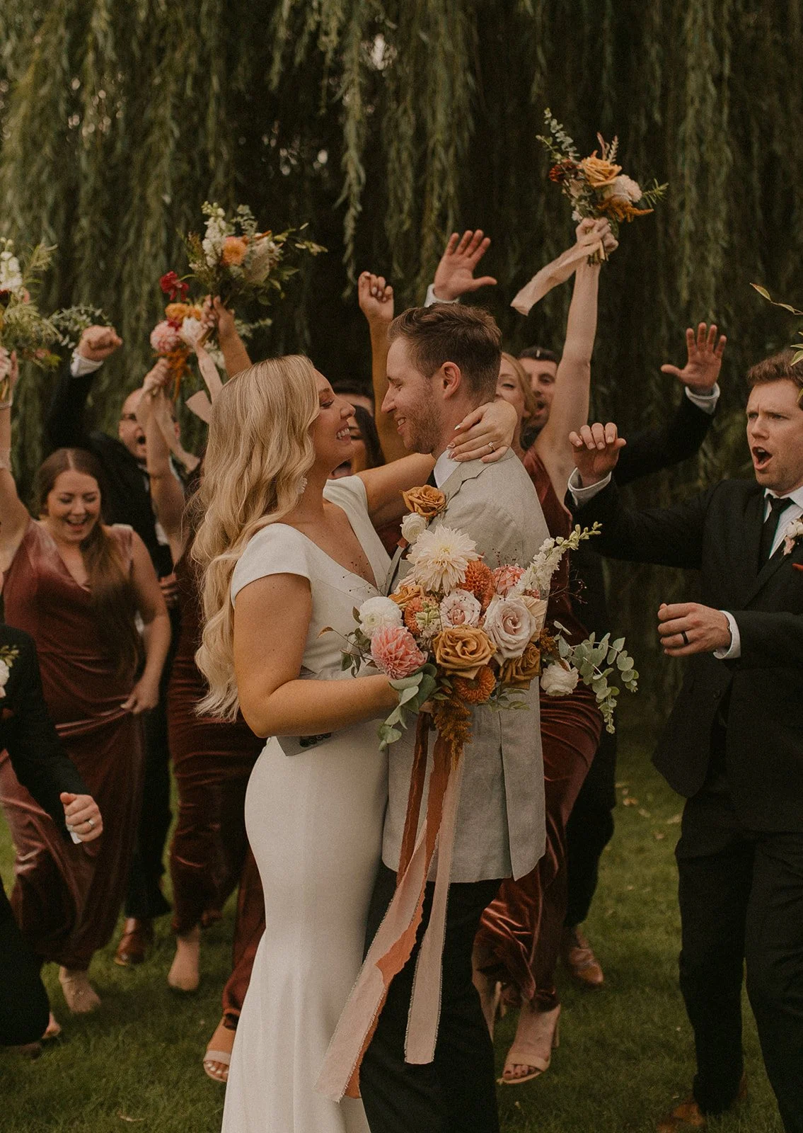 A bride and groom share a loving moment at their wedding in Northern Michigan, surrounded by joyous friends and family celebrating outdoors with flowers and festive attire.