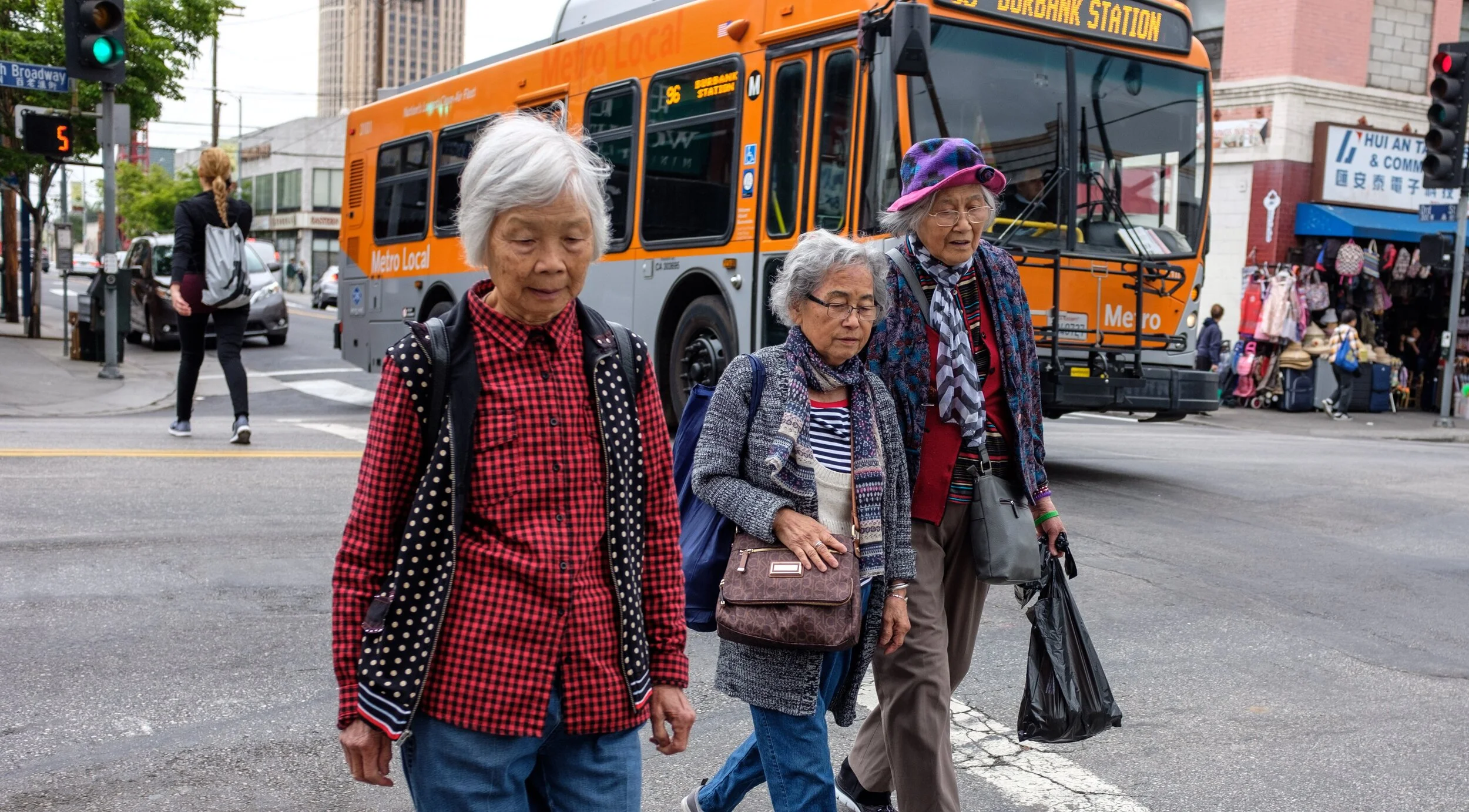 Image description:  Three Asian American senior citizens crossing a street in Chinatown with a bus in the background. Photo credit:  Rudy Espinoza