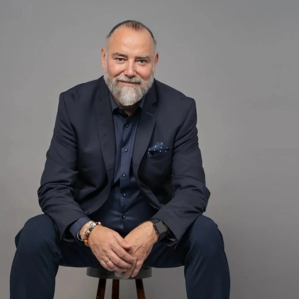 Andreas Seltmann mit grauem Bart, dunklem Anzug, blauer Hemd, weißen Turnschuhen, sitzt auf einem Holhocker vor einem grauen Hintergrund.