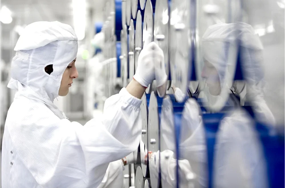 Person in glänzend weißem Schutzanzug und Maske arbeitet in einem Reinraum mit blauen Behältern, die an der Wand montiert sind.