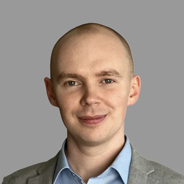 Ein Mann mit Glatze trägt einen grauen Anzug und ein blaues Hemd vor einem grauen Hintergrund.