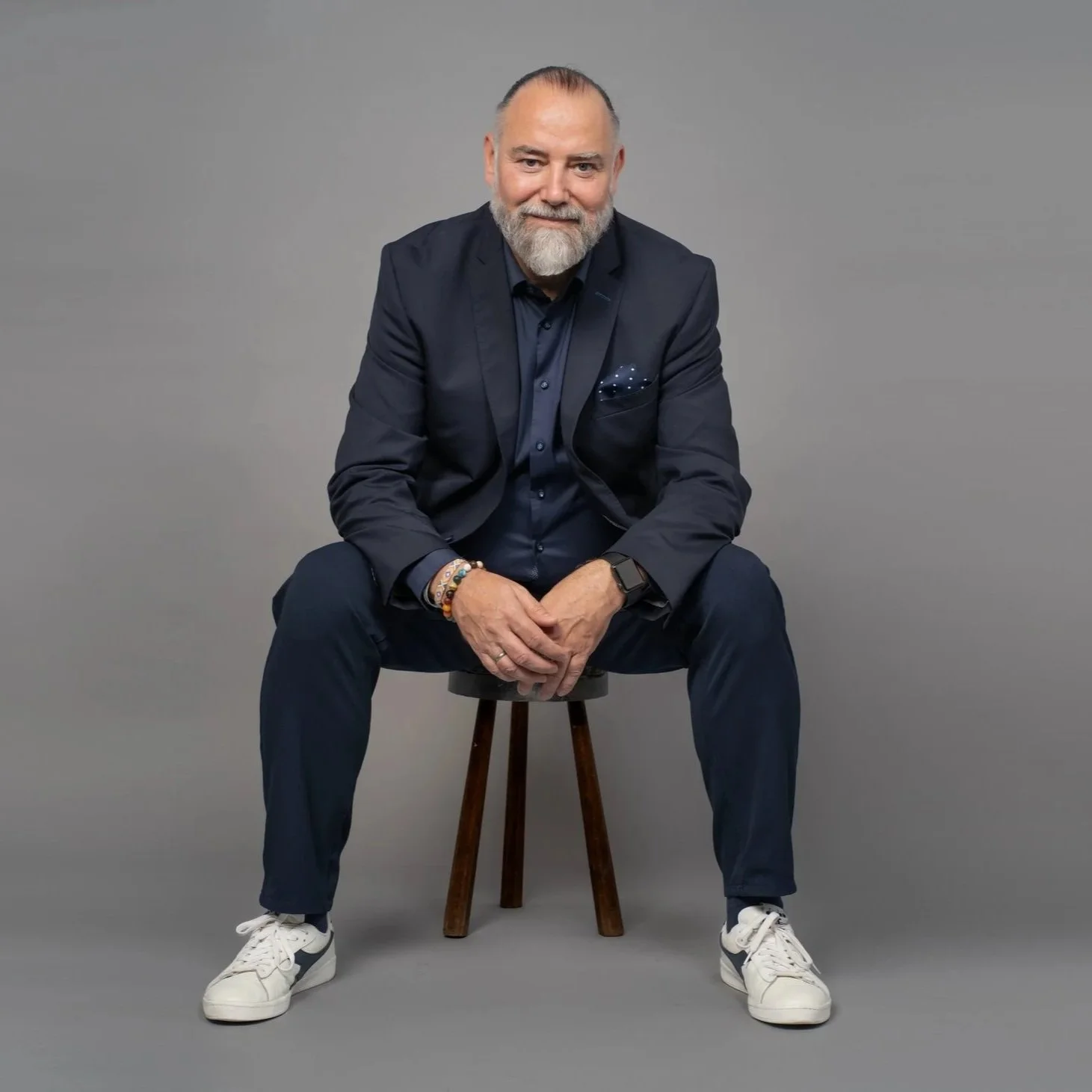 Andreas Seltmann mit grauem Bart, dunklem Anzug, blauer Hemd, weißen Turnschuhen, sitzt auf einem Holhocker vor einem grauen Hintergrund.