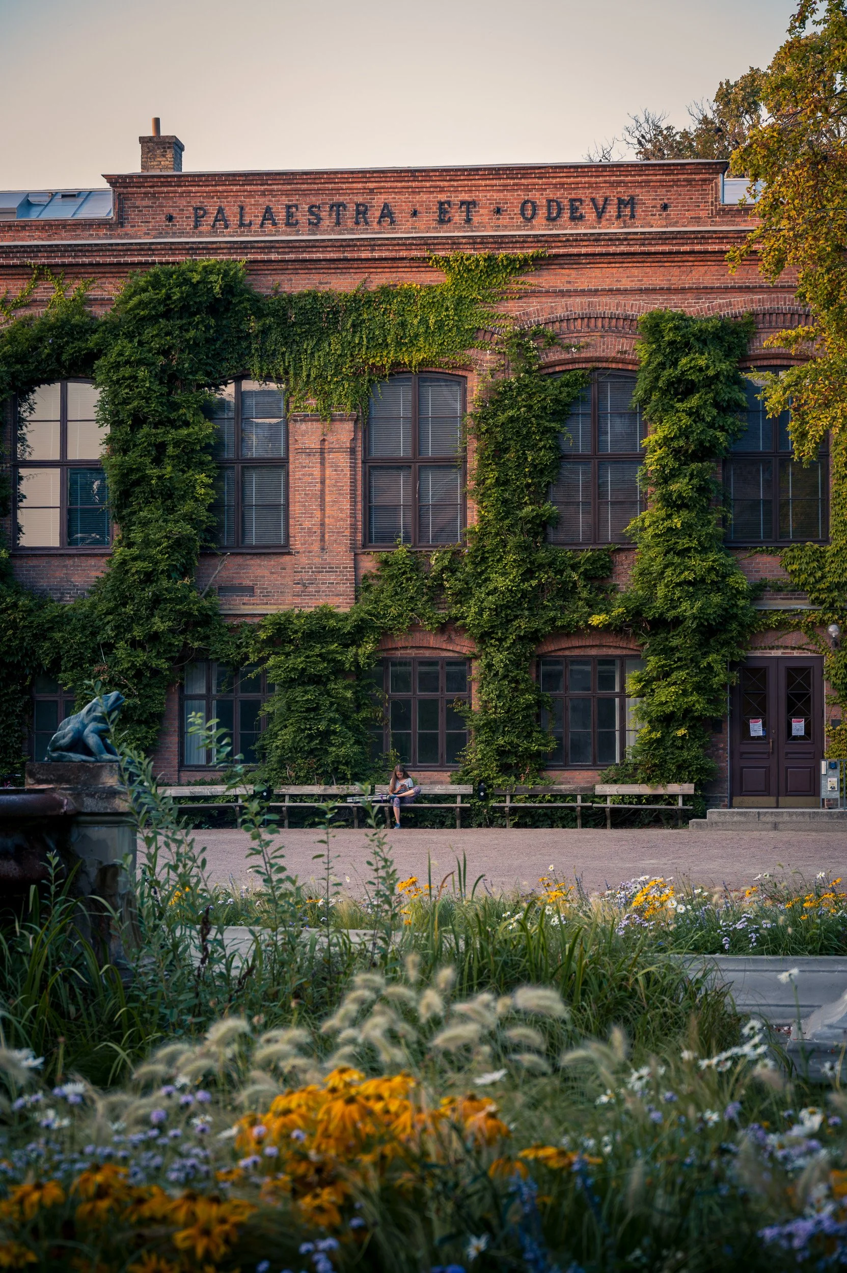 Architectural photography of Palaestra et Odeum in Lund, Sweden, featuring a historic brick facade with climbing vines behind a lush garden.