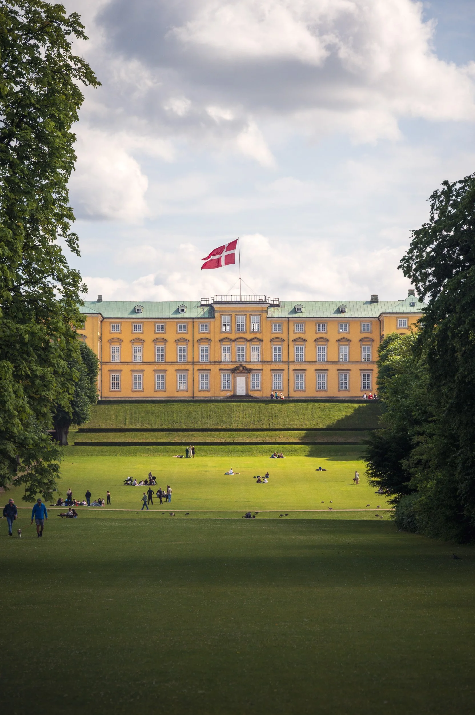 Landscape photography of the yellow Frederiksberg Palace in Copenhagen, featuring a vibrant green park lawn and the Danish flag.