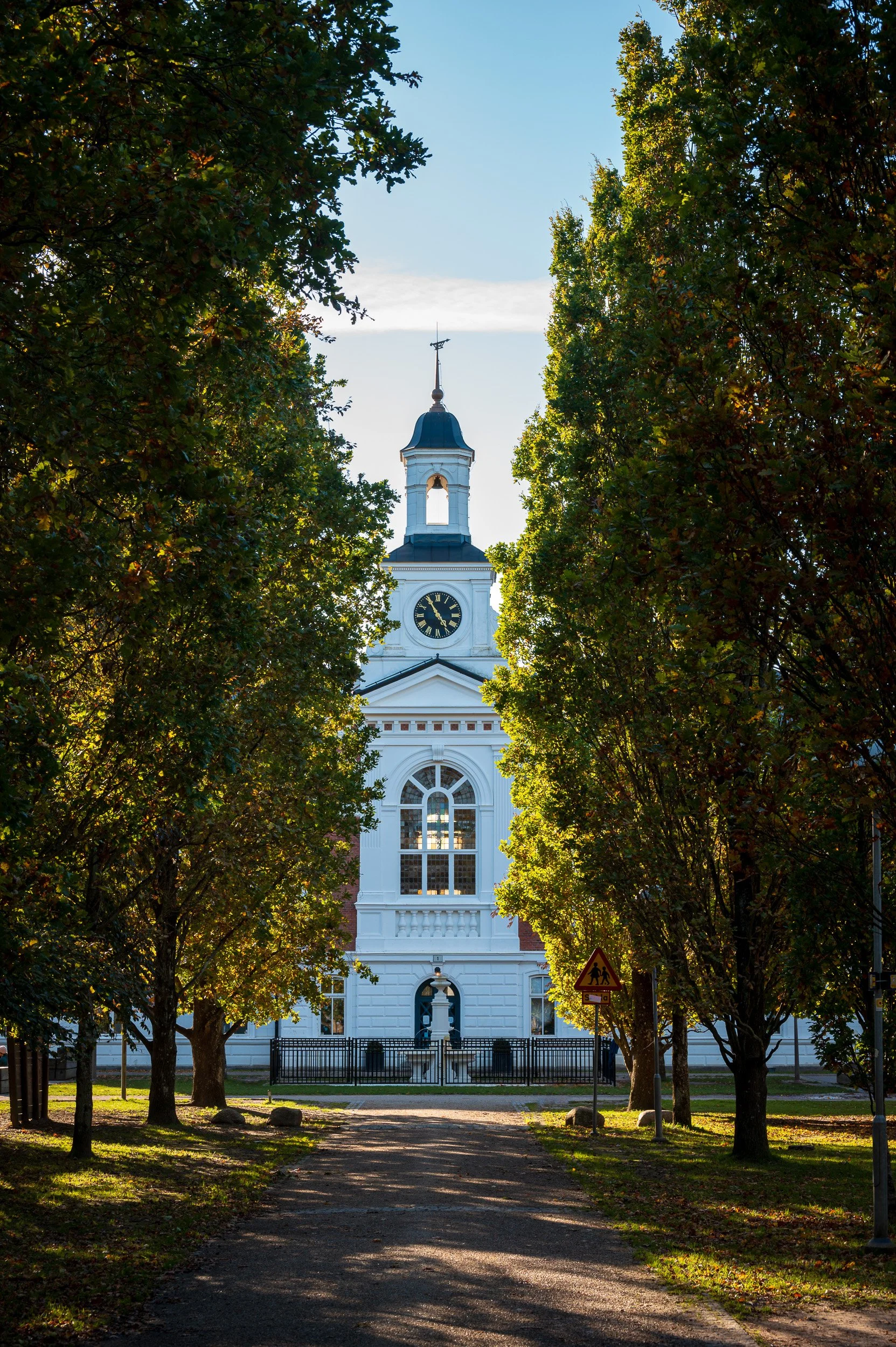 Architectural photography of a historic white building with a clock tower in the St Lars area of Lund, Sweden, framed by mature trees.