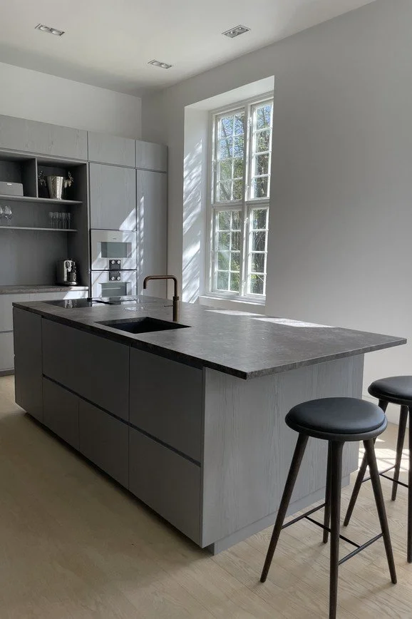 Moderne, minimalistisk køkken med gråt kabinet, sort køkkenbord, to sorte barstole og et vindue, der lader naturligt lys ind.