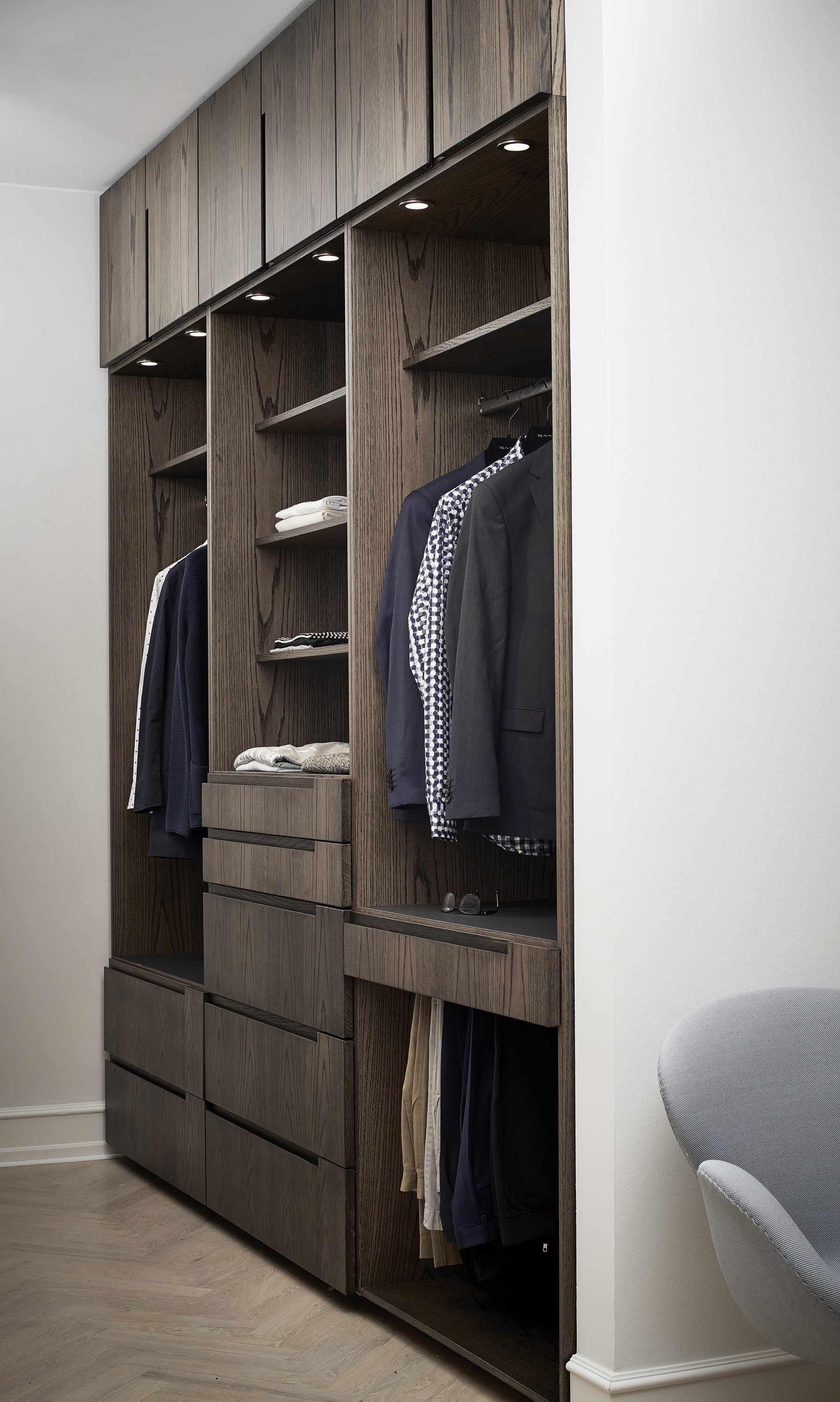 Værelse med indbygget træskab og tøj hængende, reoler med foldedetøj, solbriller og bukser på gulvet.
