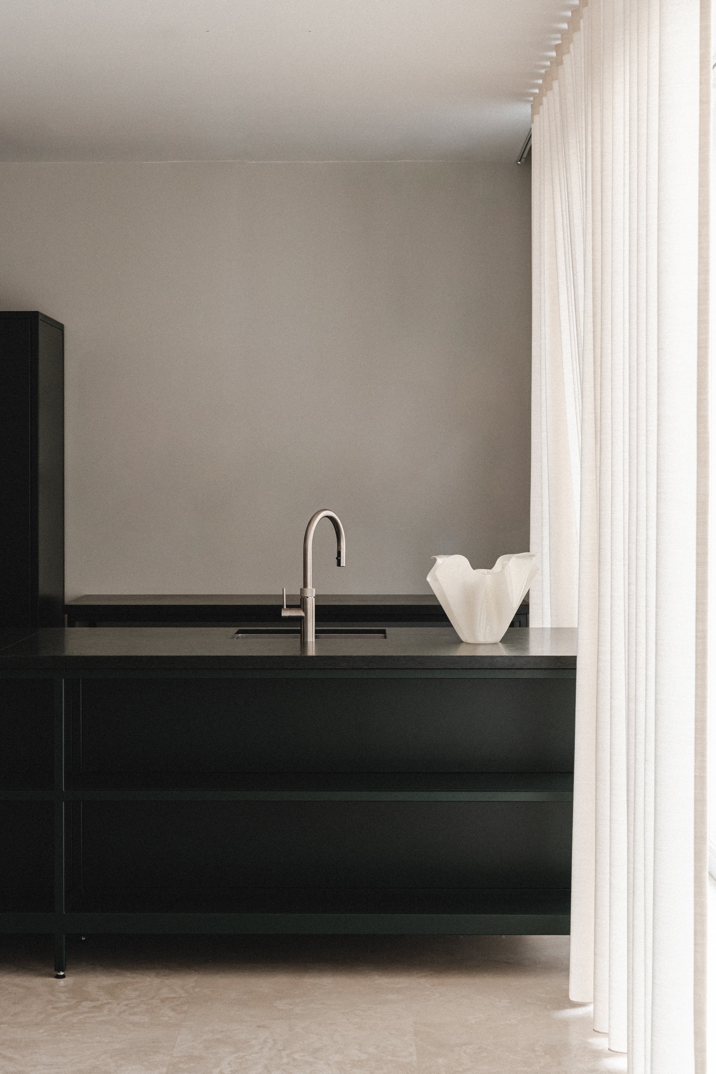 Moderne køkken med sort skab, stålvask, hvid vase på bord, og lyse gardiner.