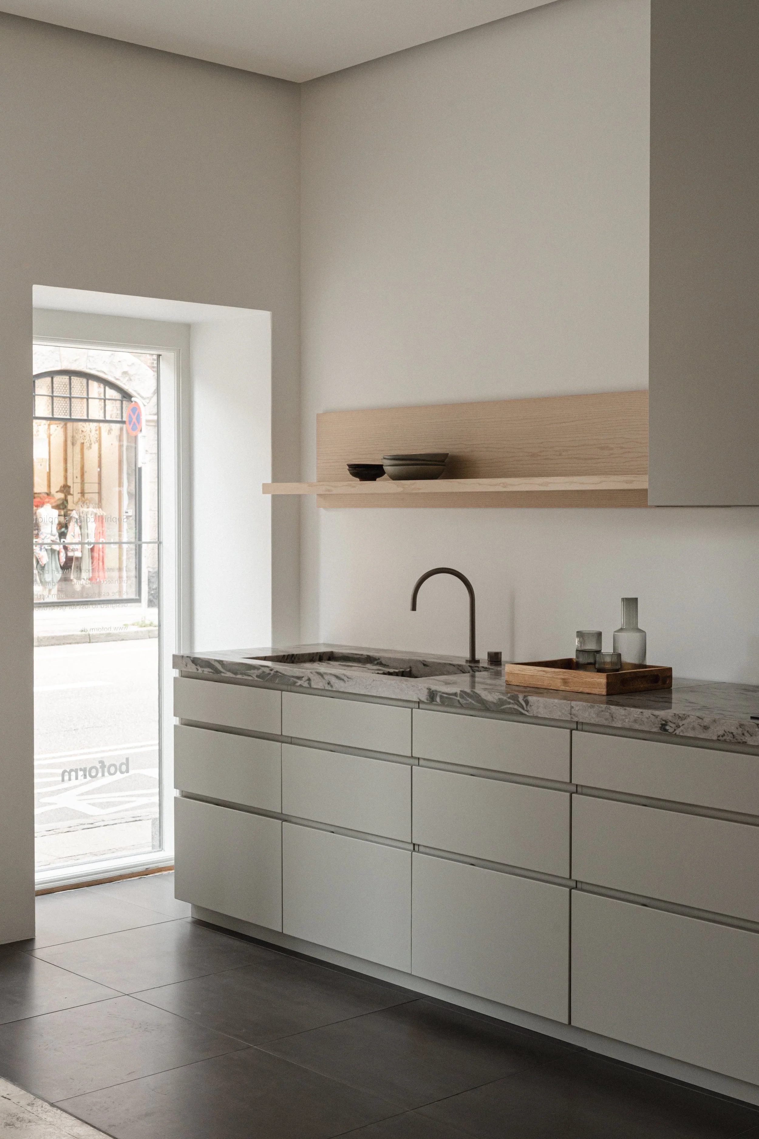 Et minimalistisk køkken med hvide skabe, grå marmorbord og en enkel vask med en sort armatur, en træhylde med skåle, og et stort vindue, der giver naturligt lys.