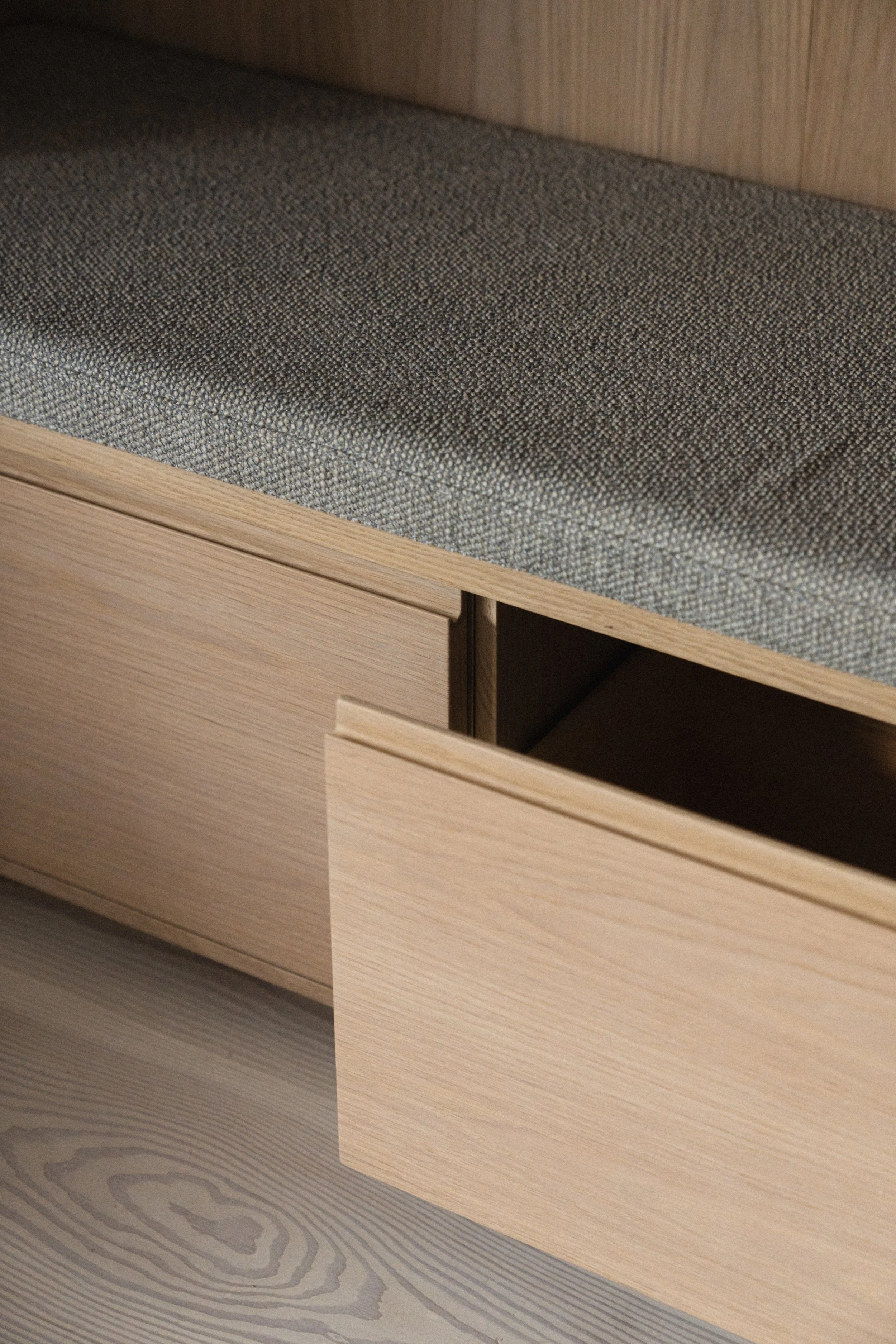 Nærbillede af en skuffe, der er halvt åben, lavet af lyst træ, med en stofsæde på en sofa eller seng ovenpå.