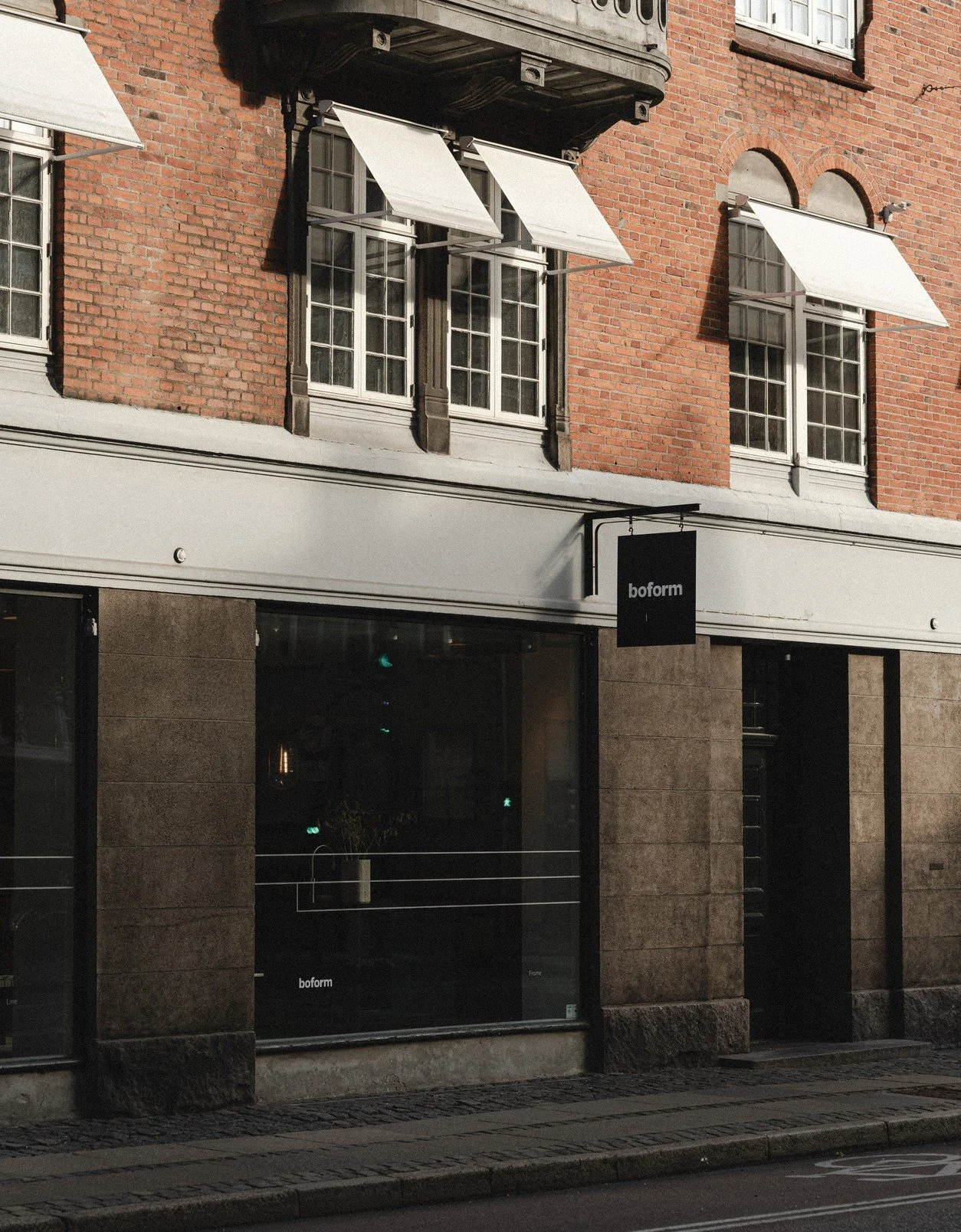 En butik kaldet 'boform' med store vinduer og en sort skiltning, beliggende i en bygning med en rød murstensfacade og hvide vinduesrammer. Der er hvide markiser over vinduerne.