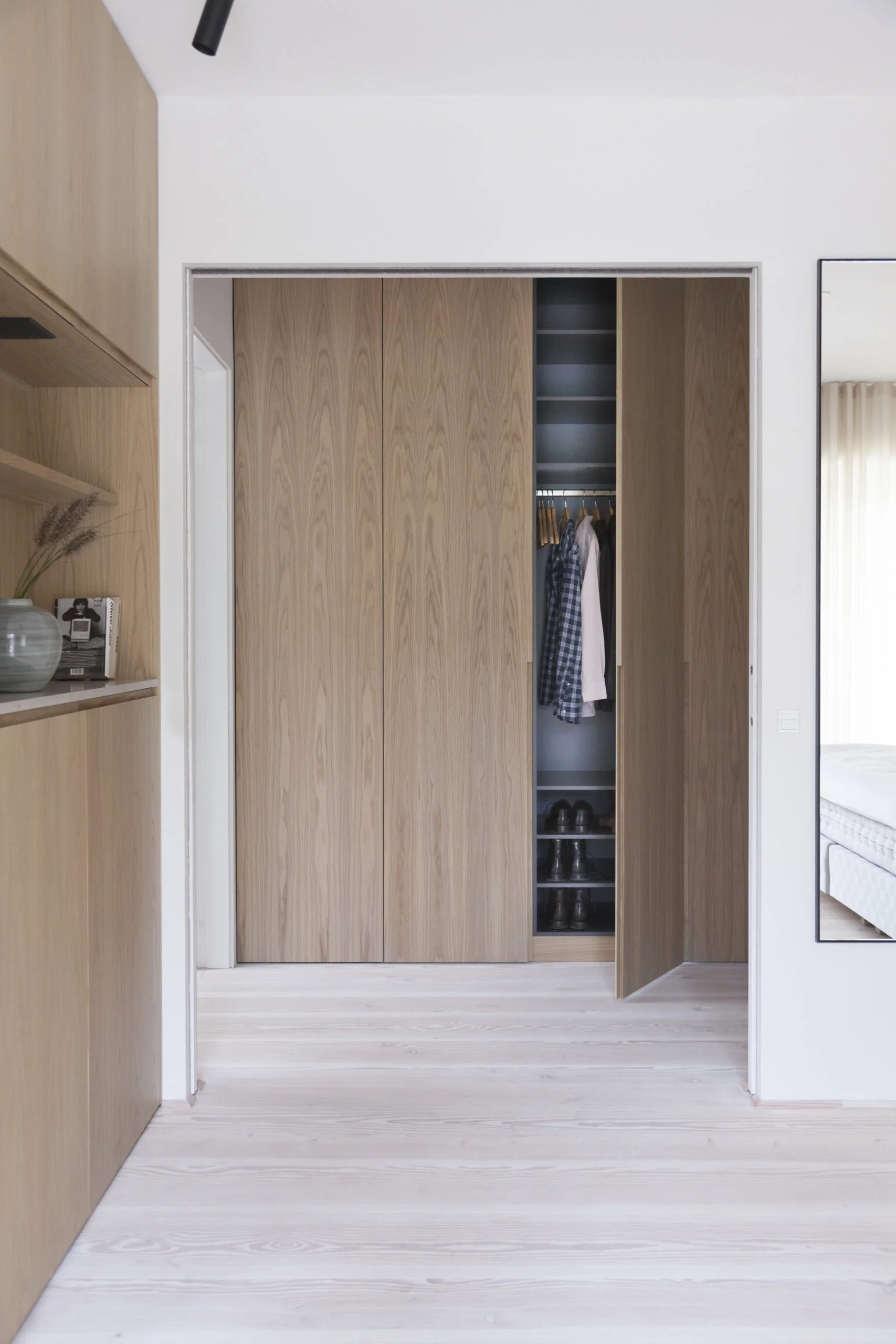 Et børnetæppe åbent skab med sko og tøj, omgivet af trædetaljer og et stort vægur. En del af et værelse med hvid væg og lyse trægulv.