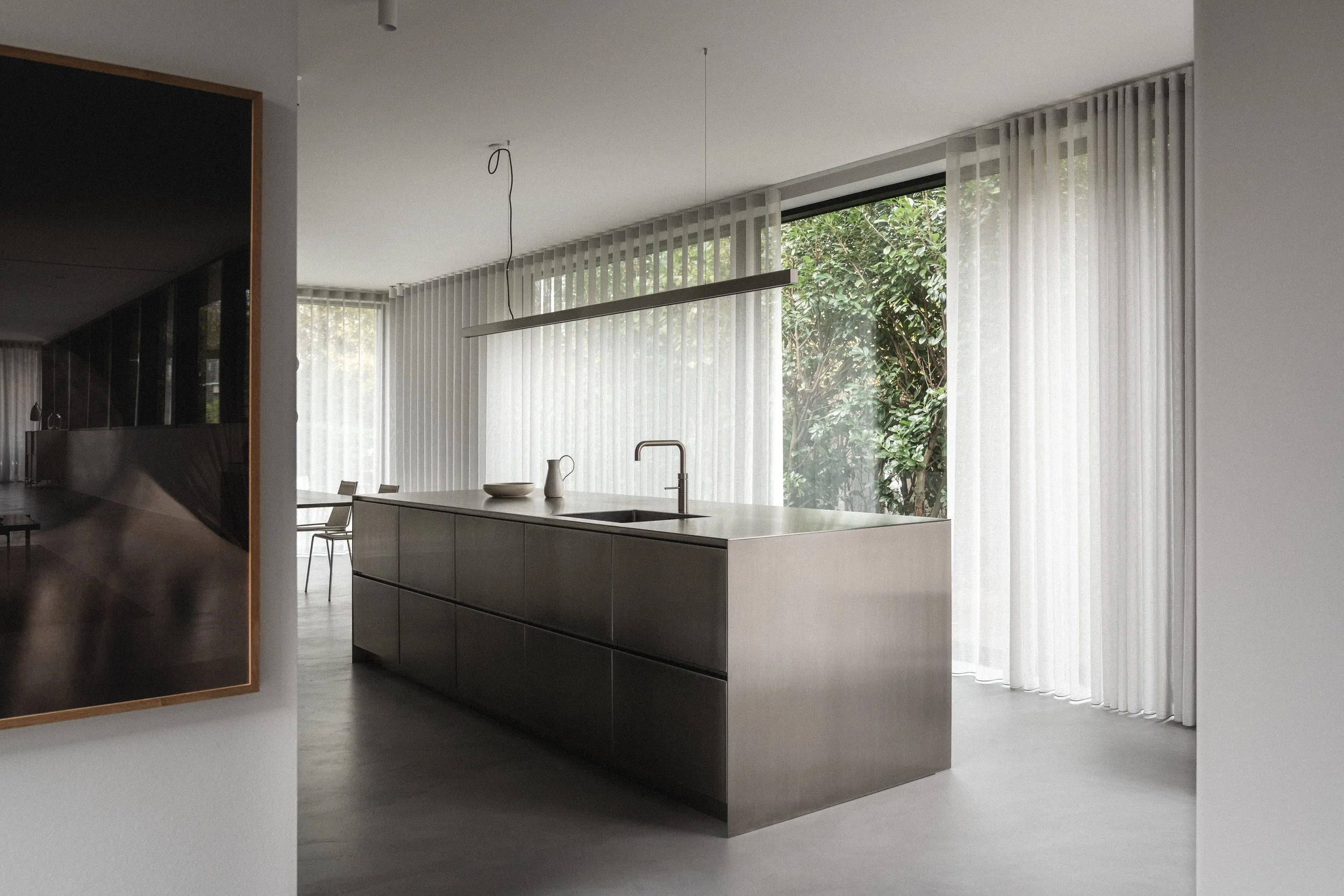 Moderne køkken med minimalistisk design, store vinduespartier med hvide lodrette blinds, enkeltbord med hvide tallerkener og kanne.