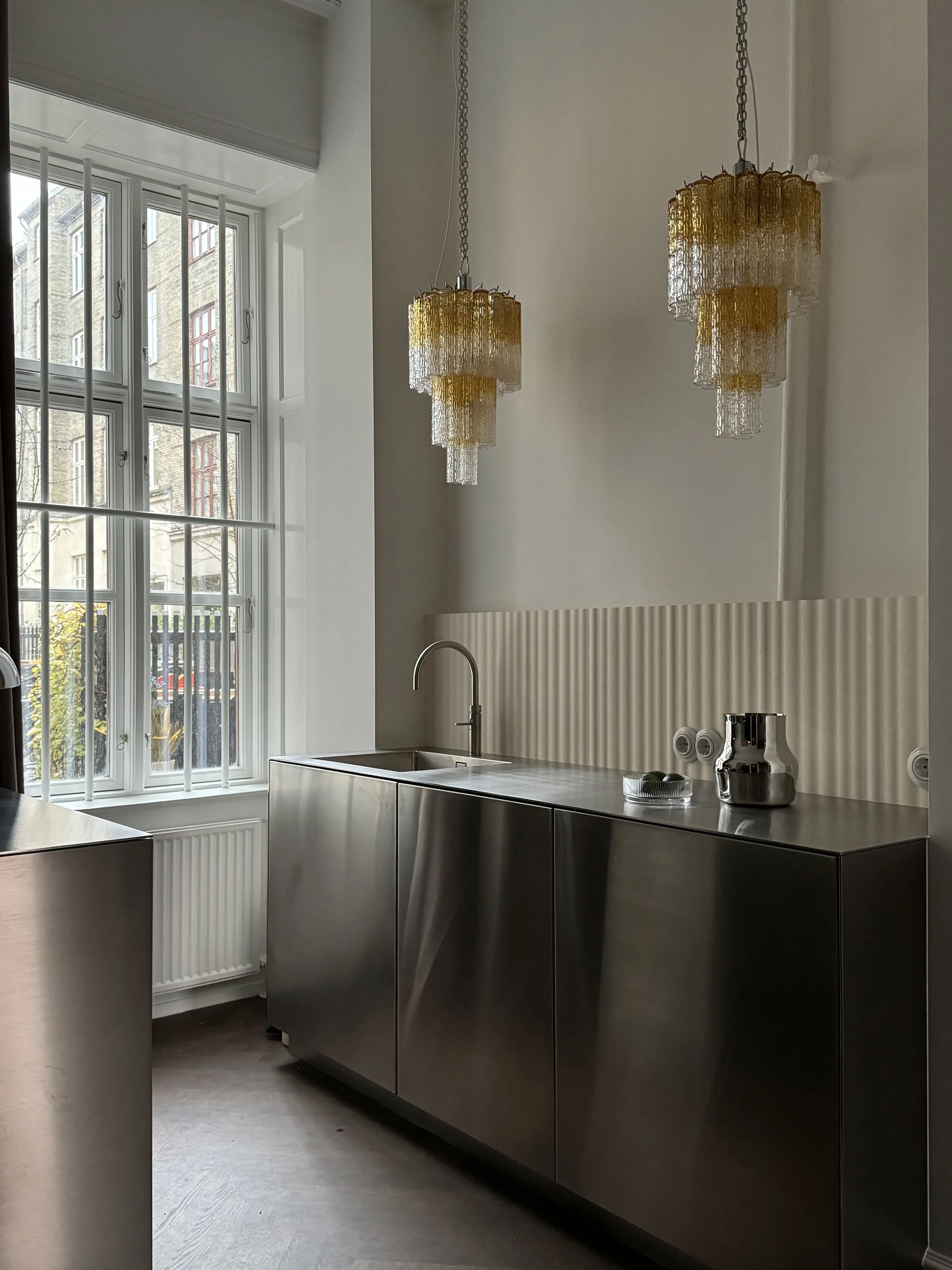 Moderne køkken med rustfrit stålskabe, stor vindue, to gulvlamper i loftet, og nogle køkkenredskaber på bordet.