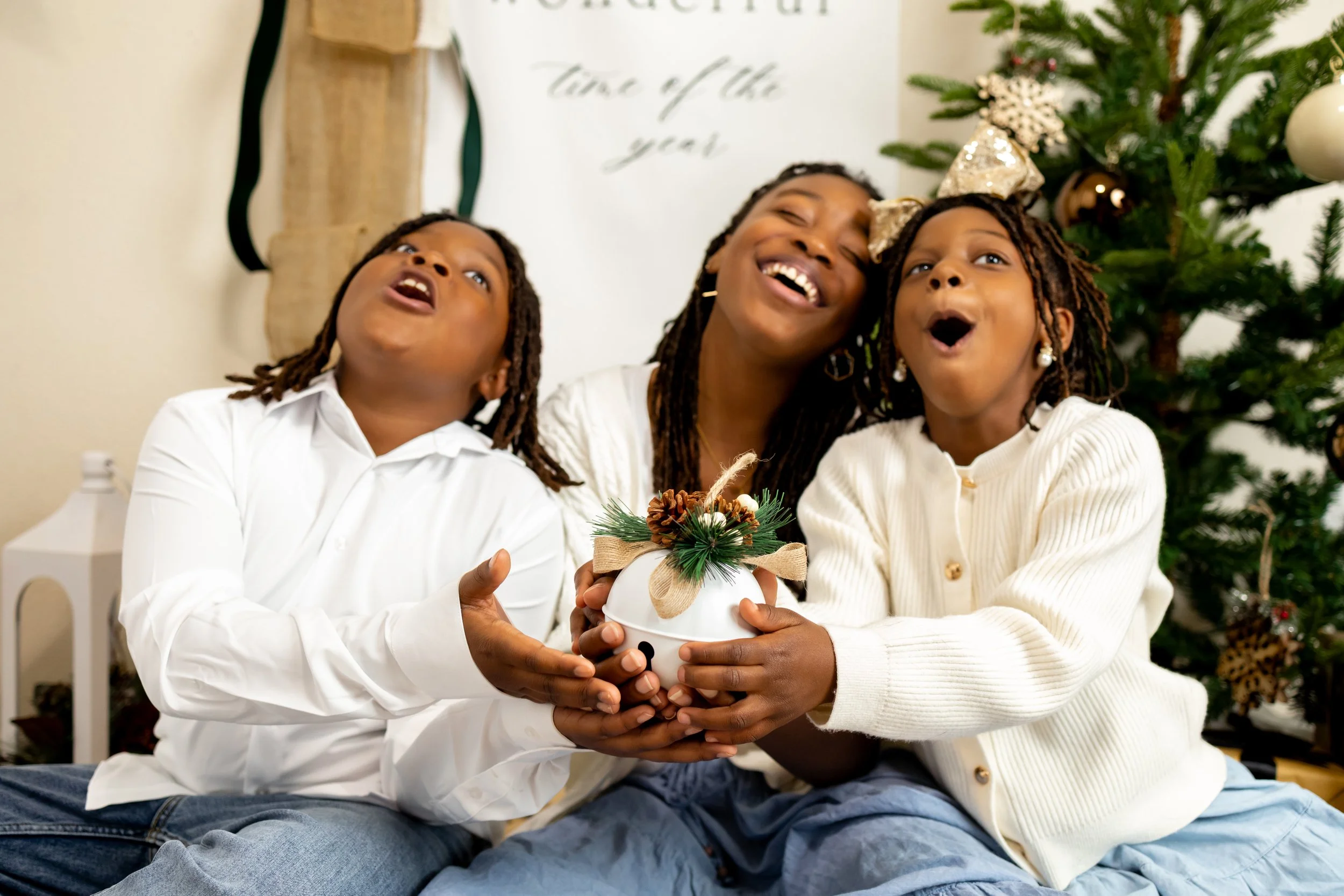 Three family members sitting in front of a Christmas tree, holding a festive holiday ornament together, appearing joyful and surprised.