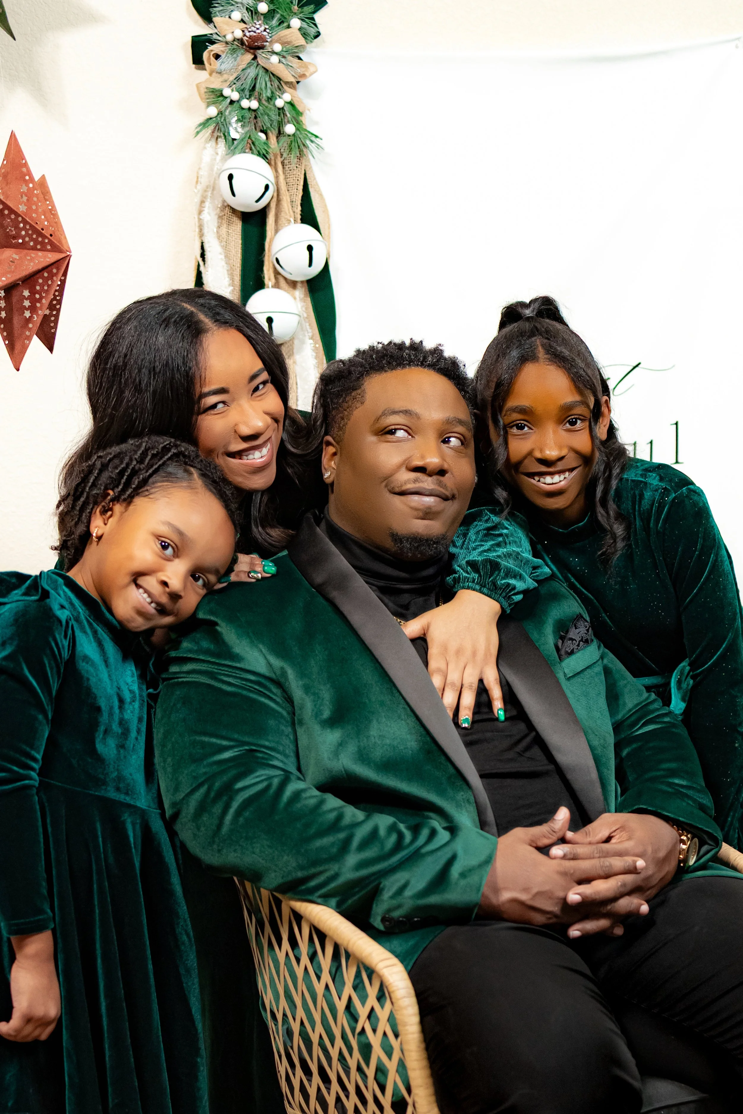 A family of five in festive green outfits posing for a holiday photo in front of Christmas decorations, including a small decorated Christmas tree on the wall.
