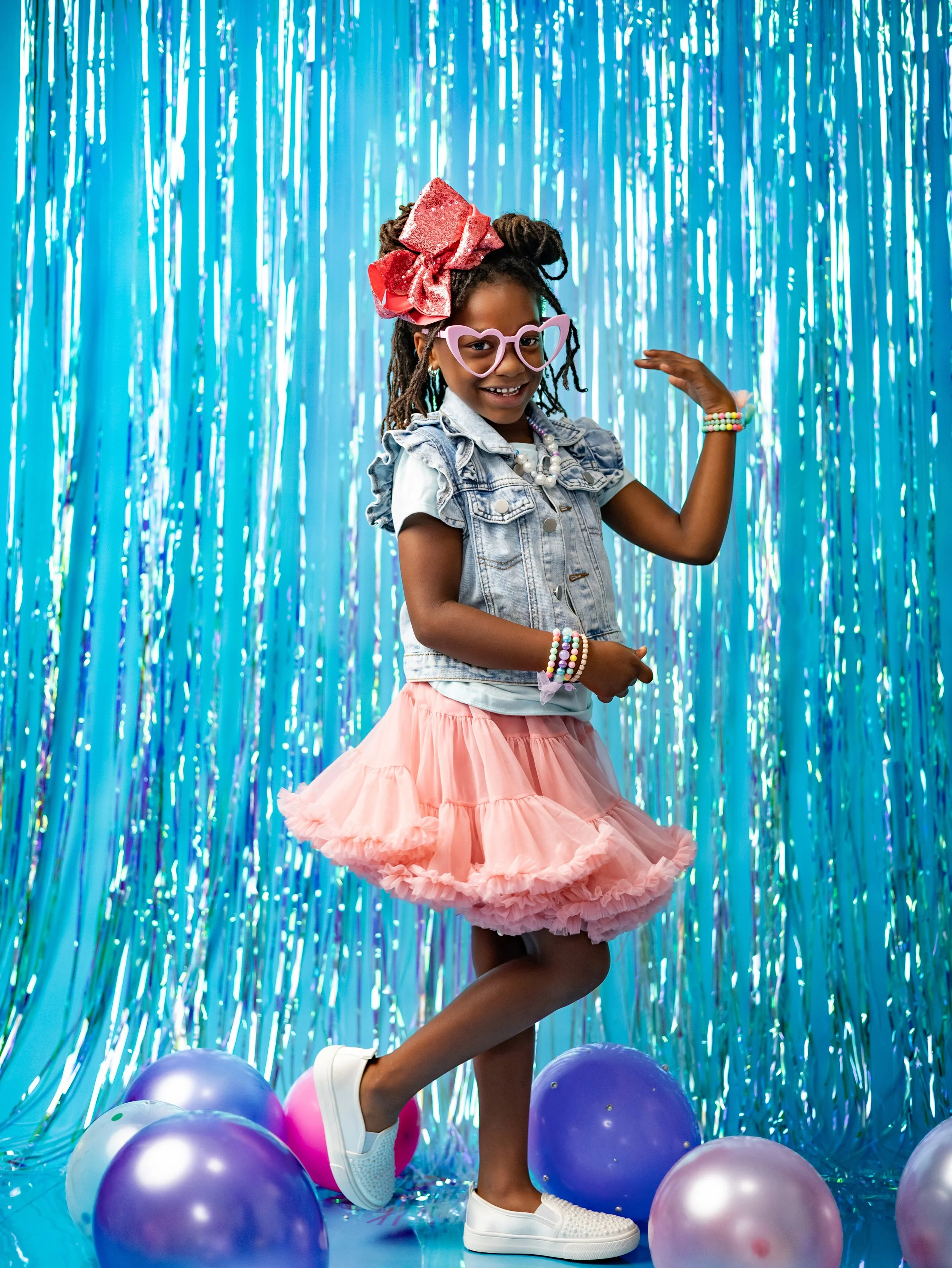 A young girl with glasses, a large bow, and jewelry is dancing among purple and pink balloons in front of a shiny blue curtain.