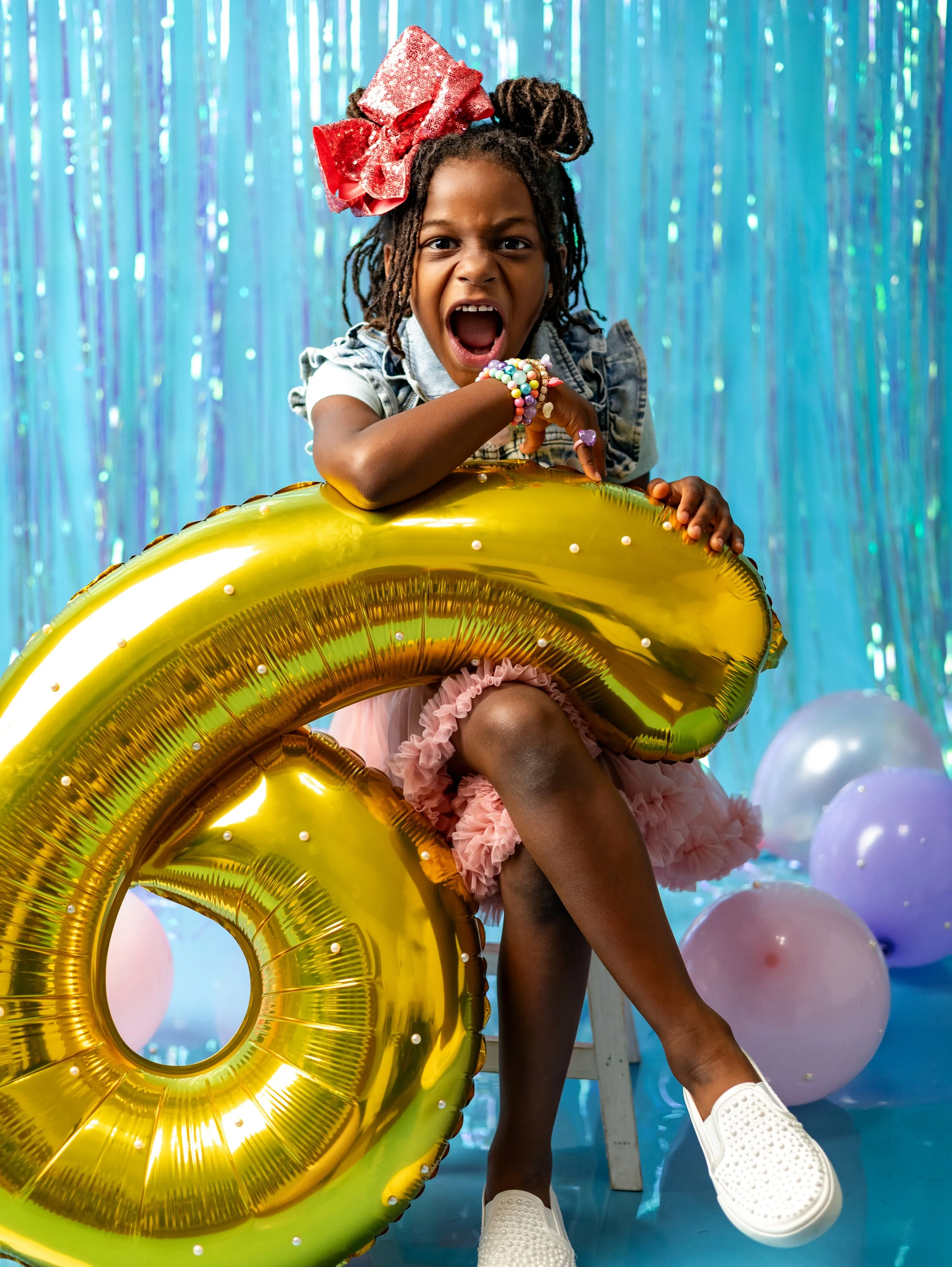Young girl with dreadlocks and a large pink bow, wearing a denim top and pink frilly skirt, sitting on a chair holding a large yellow balloon shaped like the number six, with a blue tinsel backdrop and pastel balloons around her.