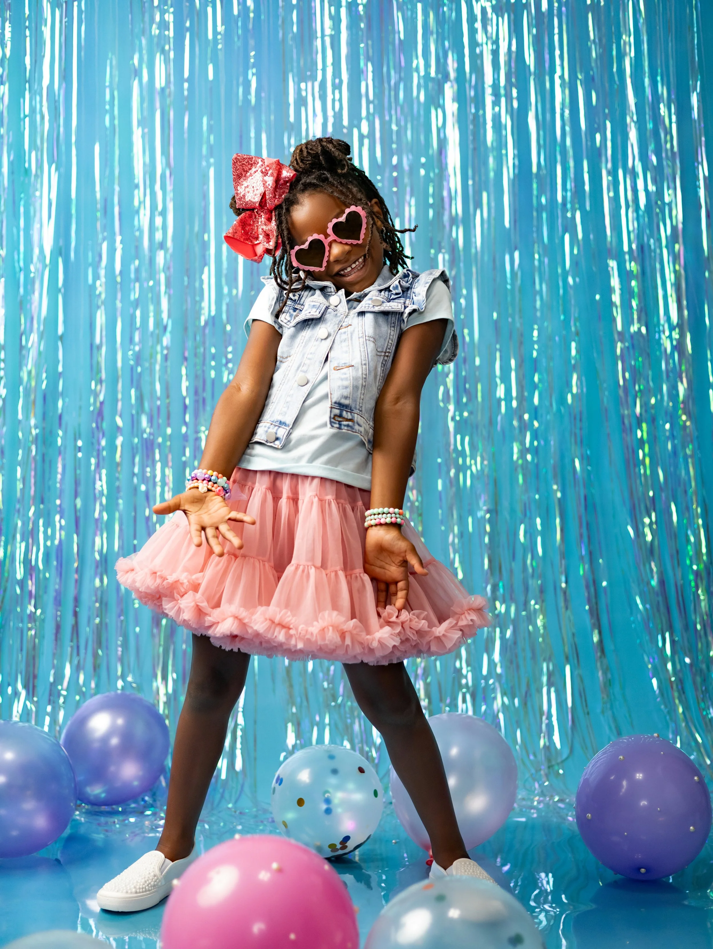 A young girl wearing heart-shaped sunglasses, a denim vest, pink tutu, and colorful bracelets, smiling and posing at a colorful party with balloons and a shiny blue streamers backdrop.