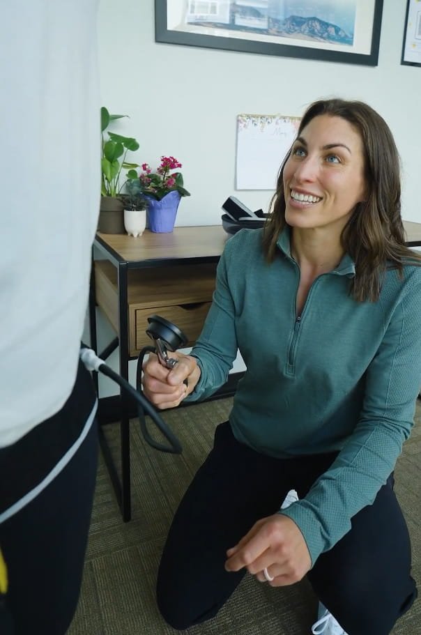 Dr. Lindsey Mayer (Boulder Sports Physiotherapy) kneeling on the floor during a medical checkup, conducting physical therapy and smiling at a patient.
