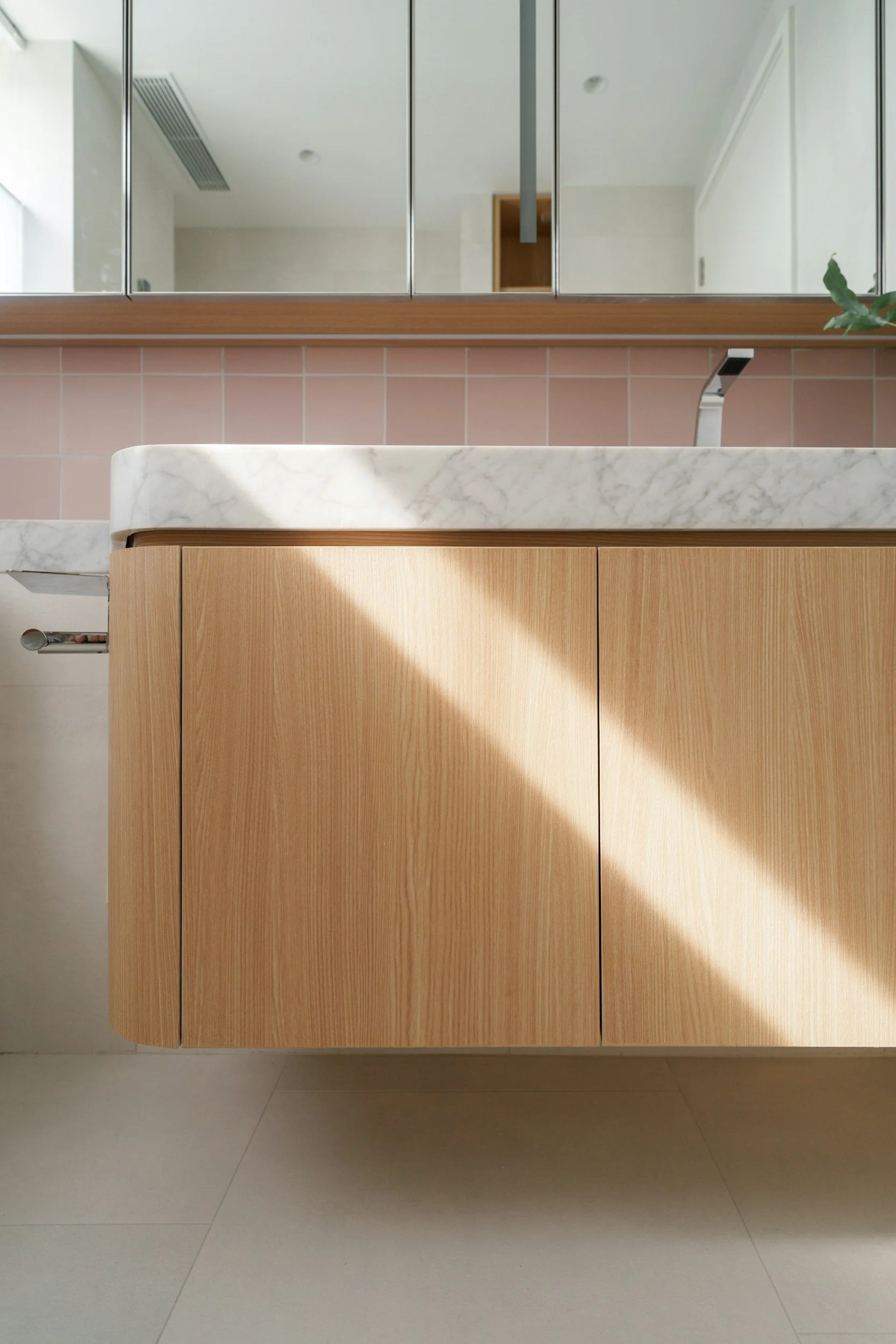 A bathroom vanity with a white marble top, wooden cabinet doors, and a black faucet. Behind the vanity is a large mirror, and the wall features pink tiles. There is a small potted plant in the top right corner of the mirror's frame.