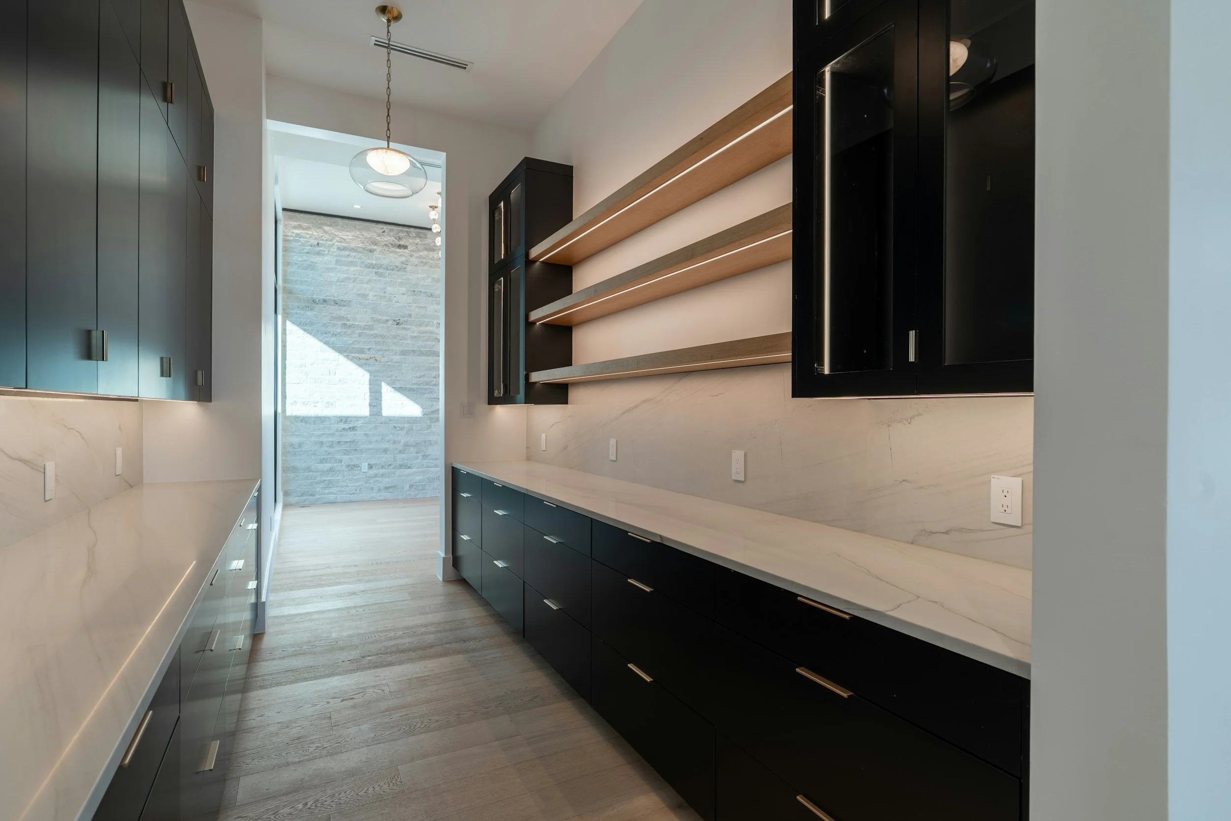 Modern kitchen with light-colored marble countertops, black lower cabinets, and black upper cabinets with glass doors. Open wooden shelves are mounted on the wall. Natural light streams in from a door with a stone wall outside, and there is a ceiling