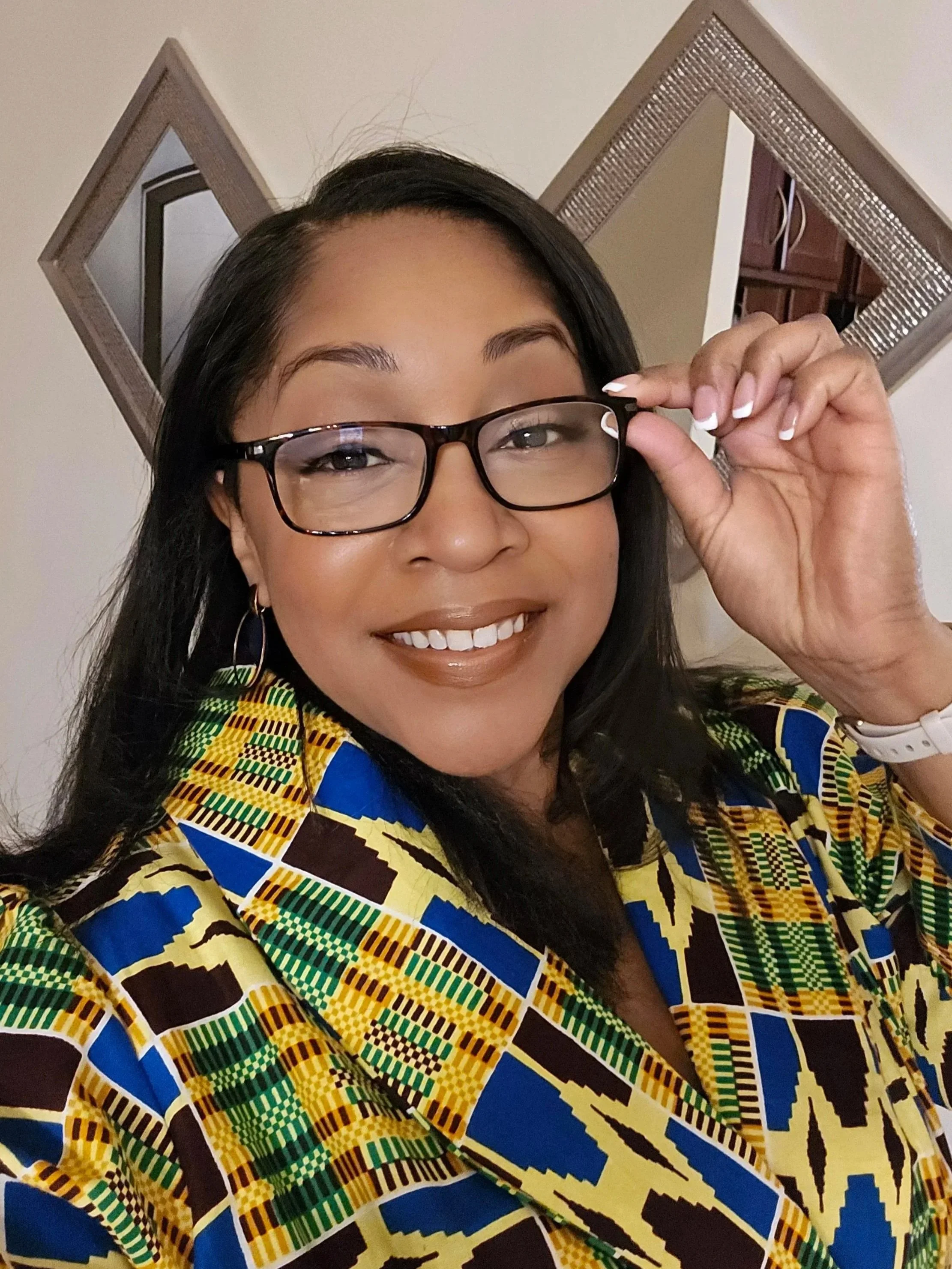 A woman with long dark hair, wearing glasses, a colorful patterned jacket, and gold hoop earrings, is smiling and holding her glasses with her right hand. Behind her are two square mirrors on a wall.