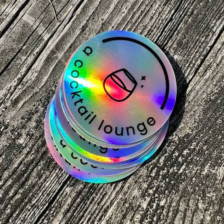 A stack of custom circular holographic stickers with black printed branding for a cocktail lounge resting on a rustic wooden surface.