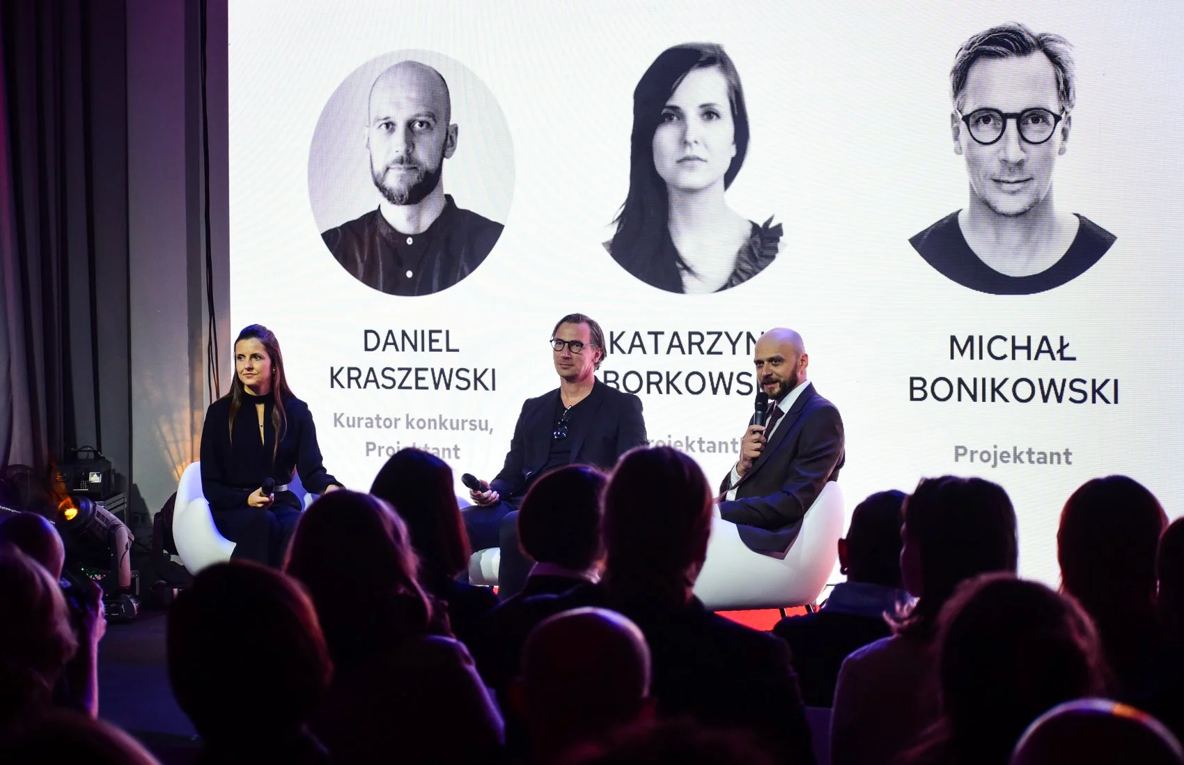 Three panelists seated on stage during a discussion at an event. Behind them is a large screen displaying black and white headshots and names of Daniel Kraszewski, Katarzyna Borkowska, and Michał Bonikowski, with their titles in Polish. The audience is visible in the foreground.